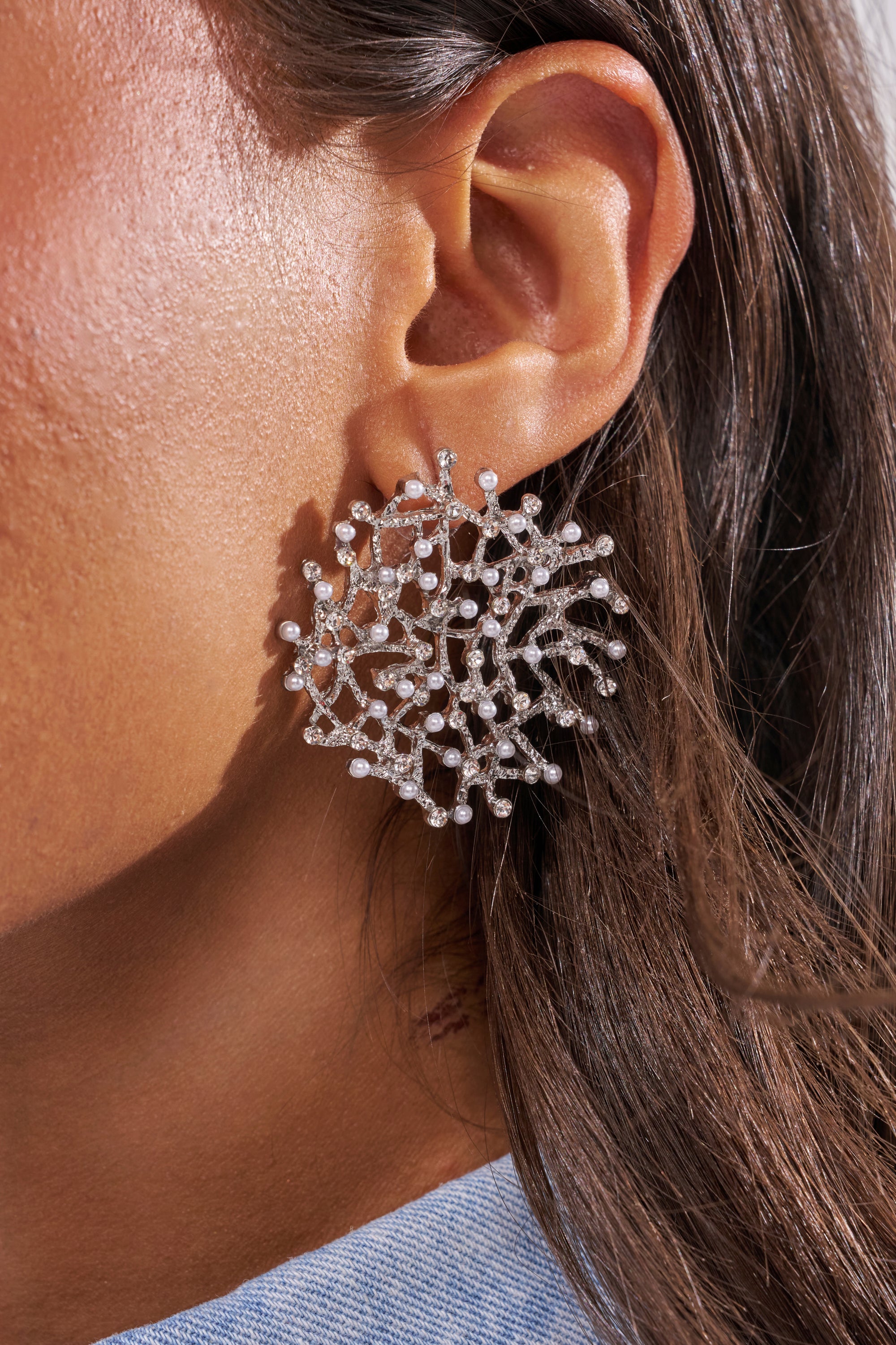 A close-up of a woman's ear highlights the TO THE REEF EARRING—an intricate silver statement piece shaped like a coral branch with pearl-like accents. Her dark hair partially covers her ear, emphasizing the earring's unique elegance. 2