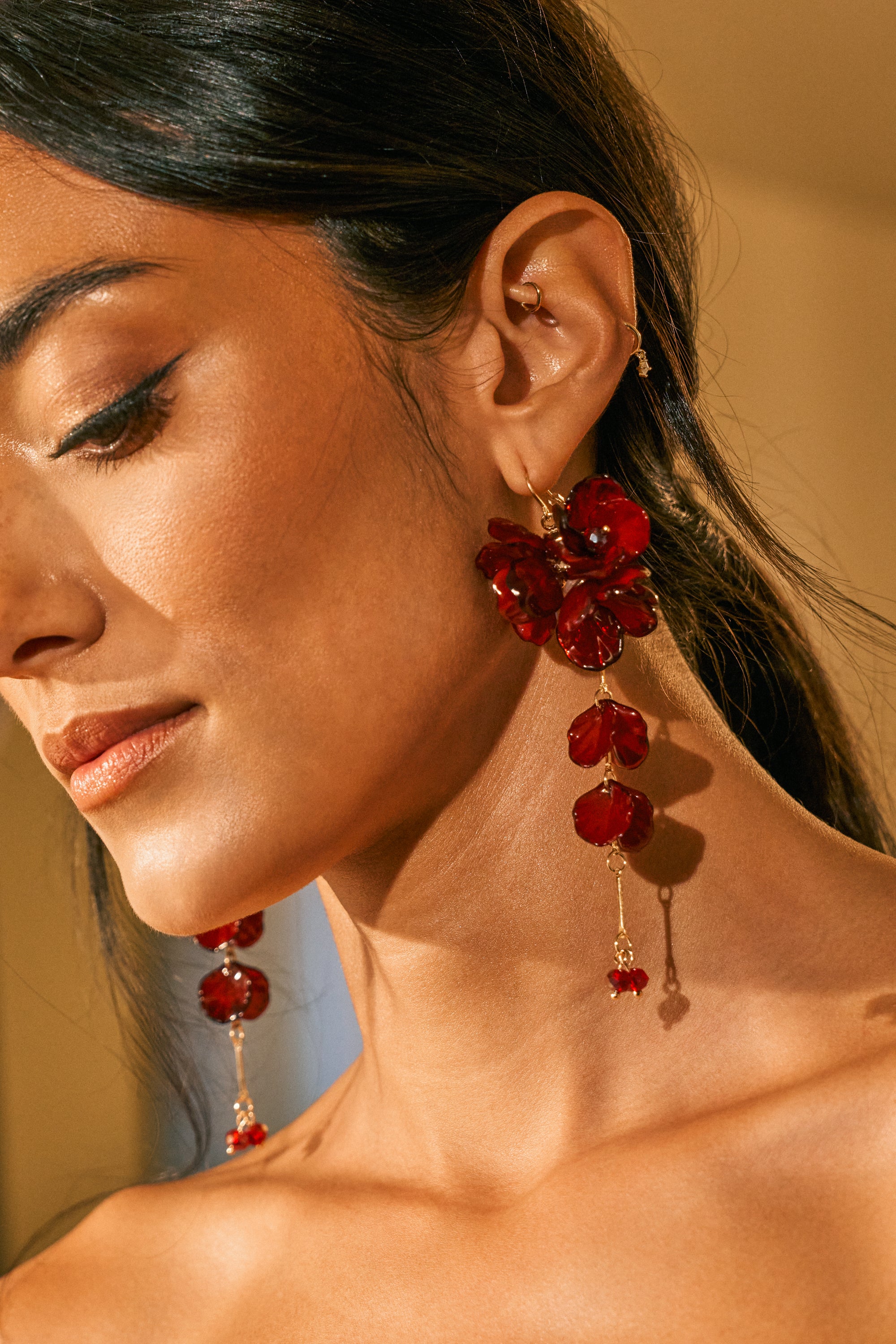 A woman with smooth skin and winged eyeliner wears the DRIP DRIP BURGUNDY DANGLE EARRING—perfect date night jewelry. Her dark hair is pulled back, showing her profile and bare shoulders in warm light.