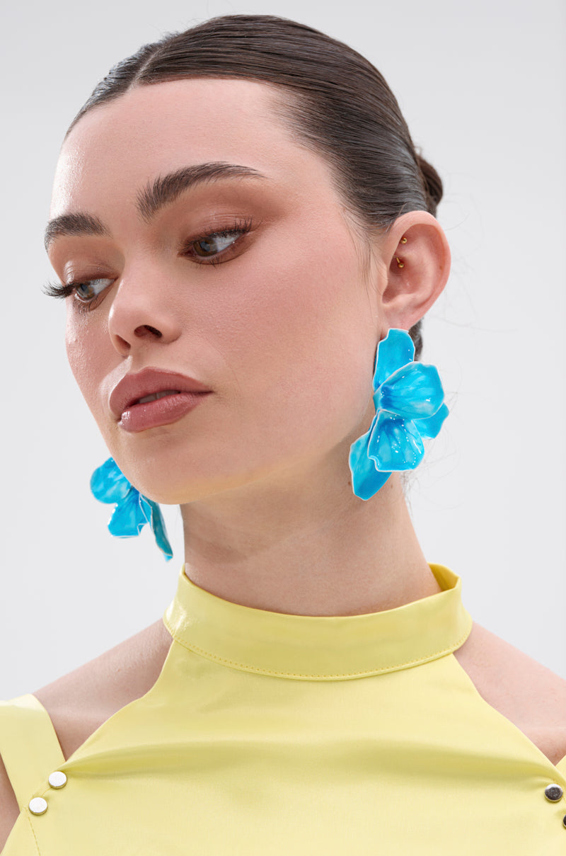 A woman with brown hair in a sleek bun wears the bright blue NO BLUES EARRING and a sleeveless yellow high-neck AKIRA Label top, looking slightly down with a neutral expression against a plain background. 1