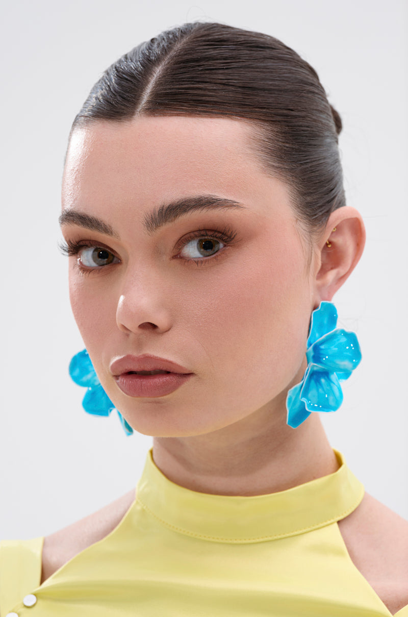 A woman with dark hair in a sleek bun models the NO BLUES EARRING, which has a bold floral design, paired with a textured light yellow high-neck top, posing against a plain light background. 4