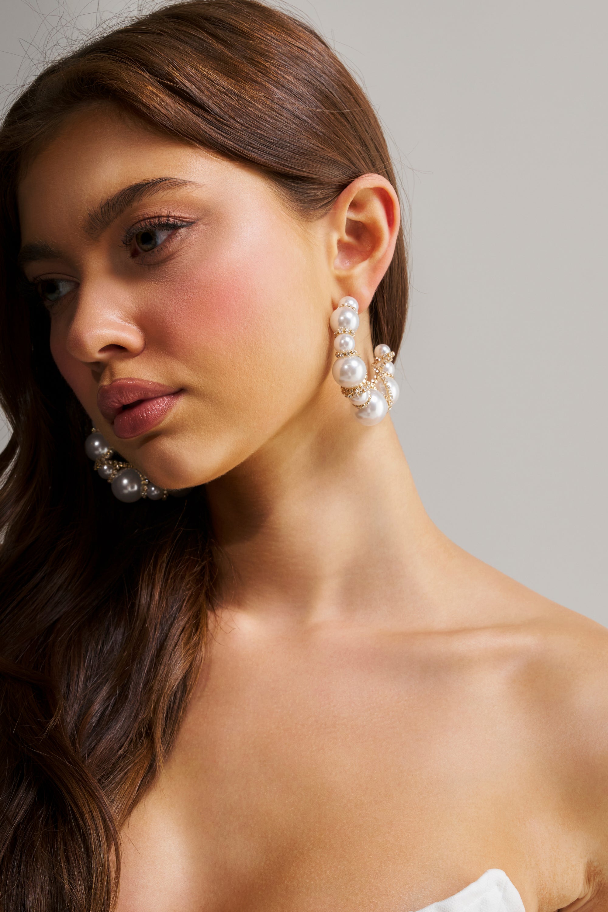 A woman with long brown hair wears the MIRACLE HOOP EARRING—large hoops adorned with white and cream pearls—paired with natural makeup and pink blush, posing against a plain light background.