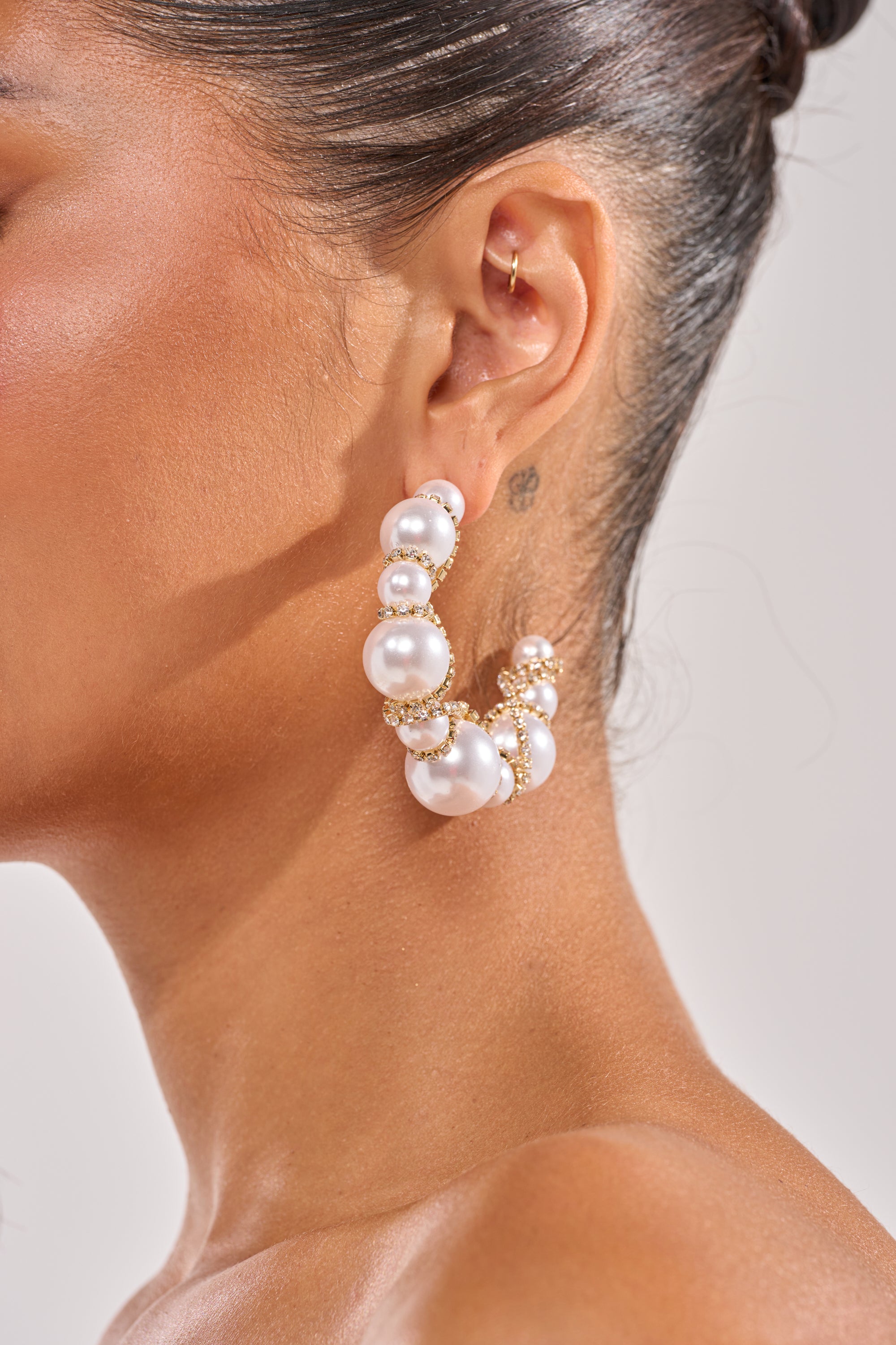 Close-up of a woman's ear wearing the MIRACLE HOOP EARRING, gold-tone hoops embellished with white pearl clusters and elegant gold accents. Her hair is pulled back, revealing a small tattoo behind her ear. 2
