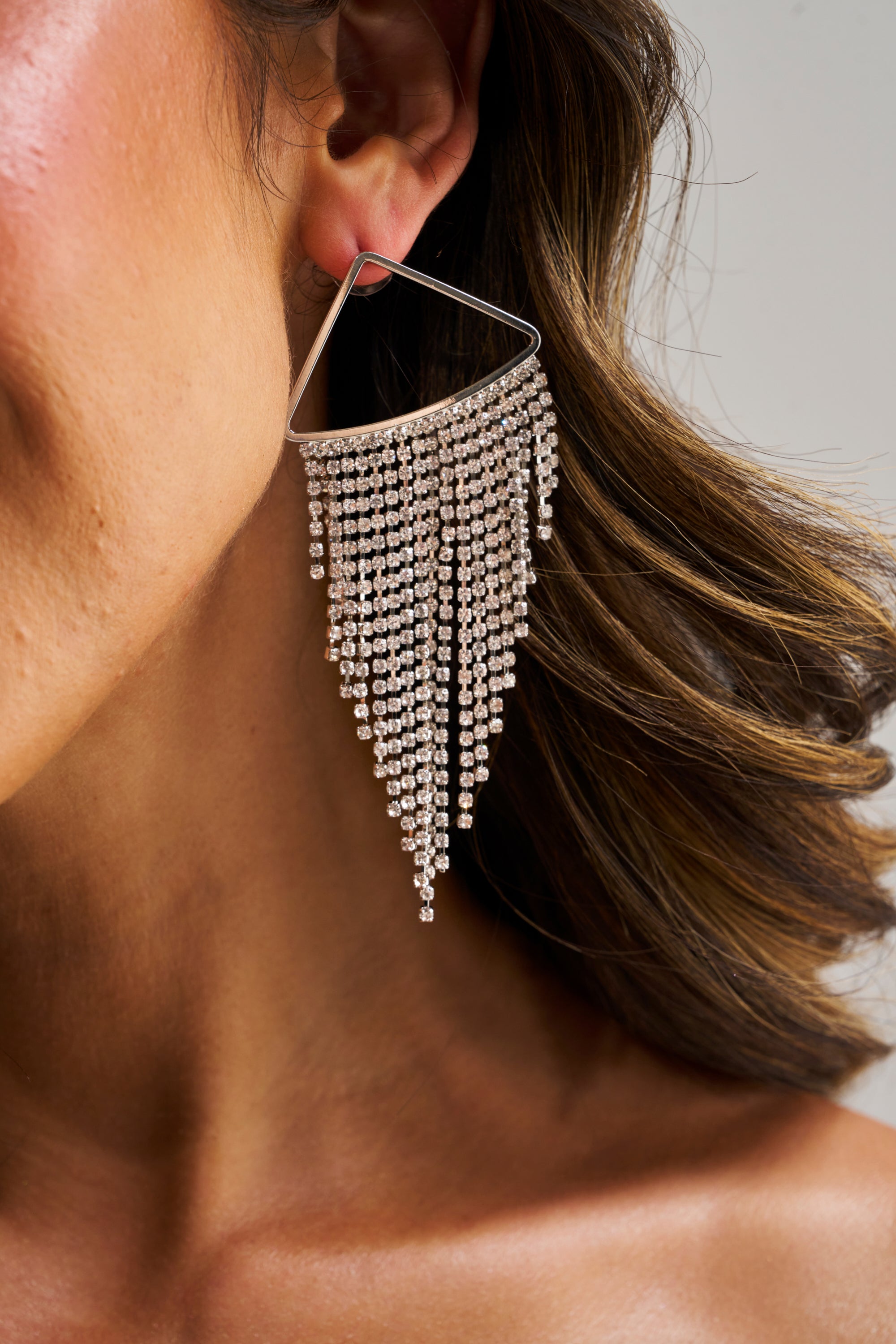 Close-up of a woman's ear wearing the WE'RE GOING OUT EARRING IN SILVER—large, triangular drop earrings with cascading rhinestones for a chandelier effect—partially framed by her wavy brown hair.