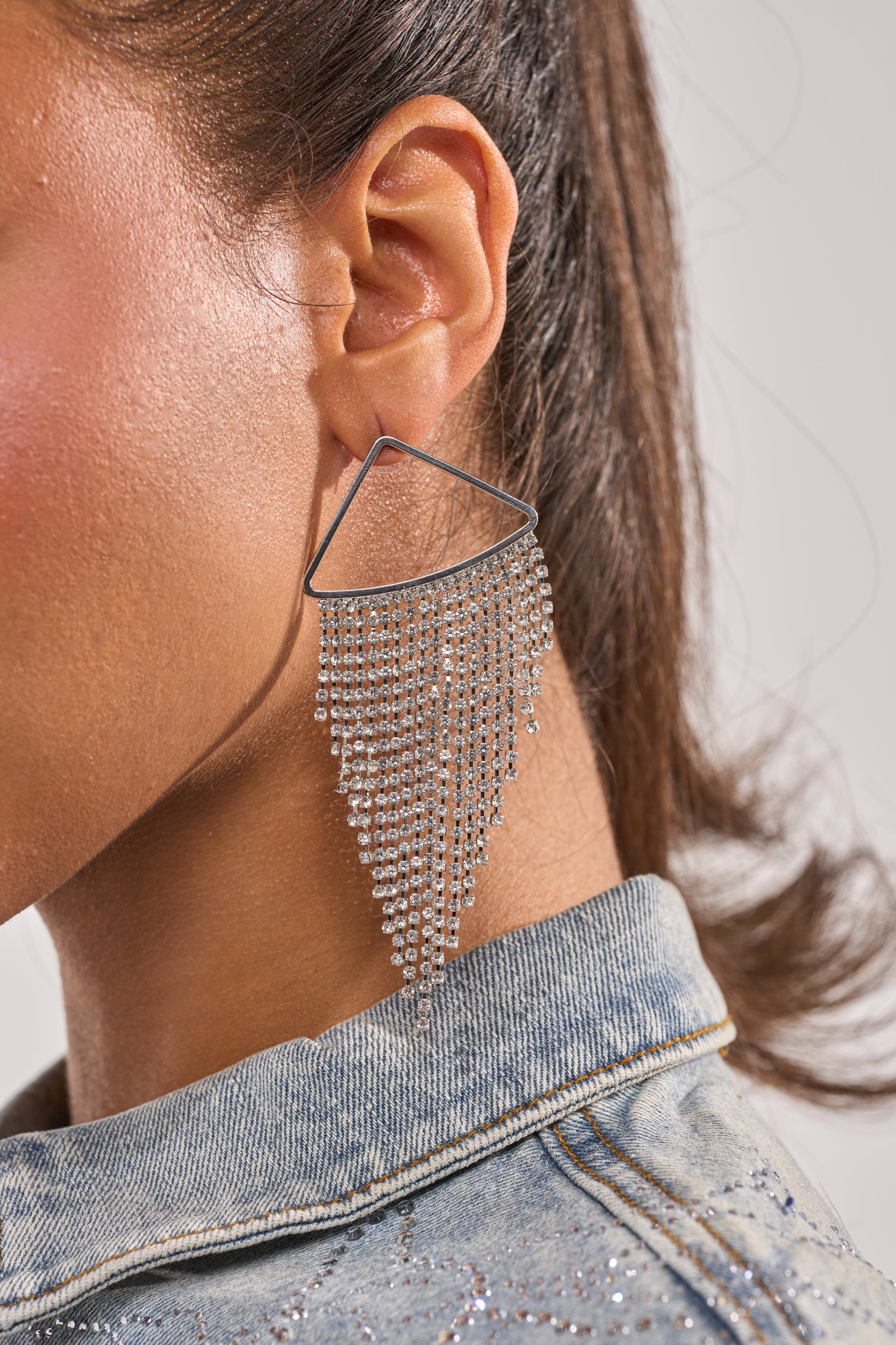 Close-up of a person with brown hair in a light blue denim jacket, wearing the WE'RE GOING OUT EARRING IN SILVER—a large, triangular chandelier design with cascading strands of small, sparkling beads. 2