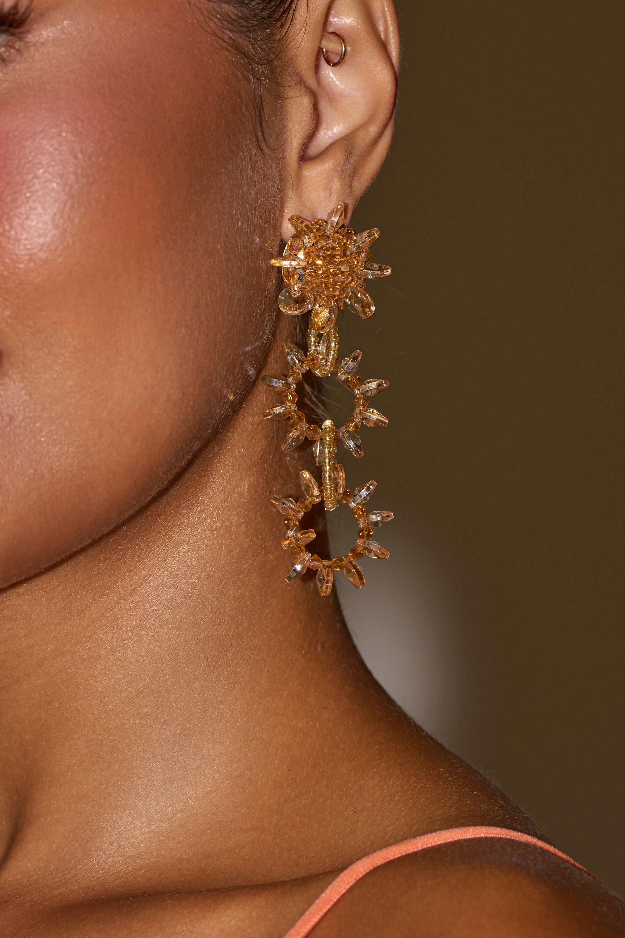 Close-up of a person with medium brown skin wearing the COME AND GET ME EARRING—large, gold, sunburst-shaped festival jewelry—paired with a peach top and an out-of-focus background. 0