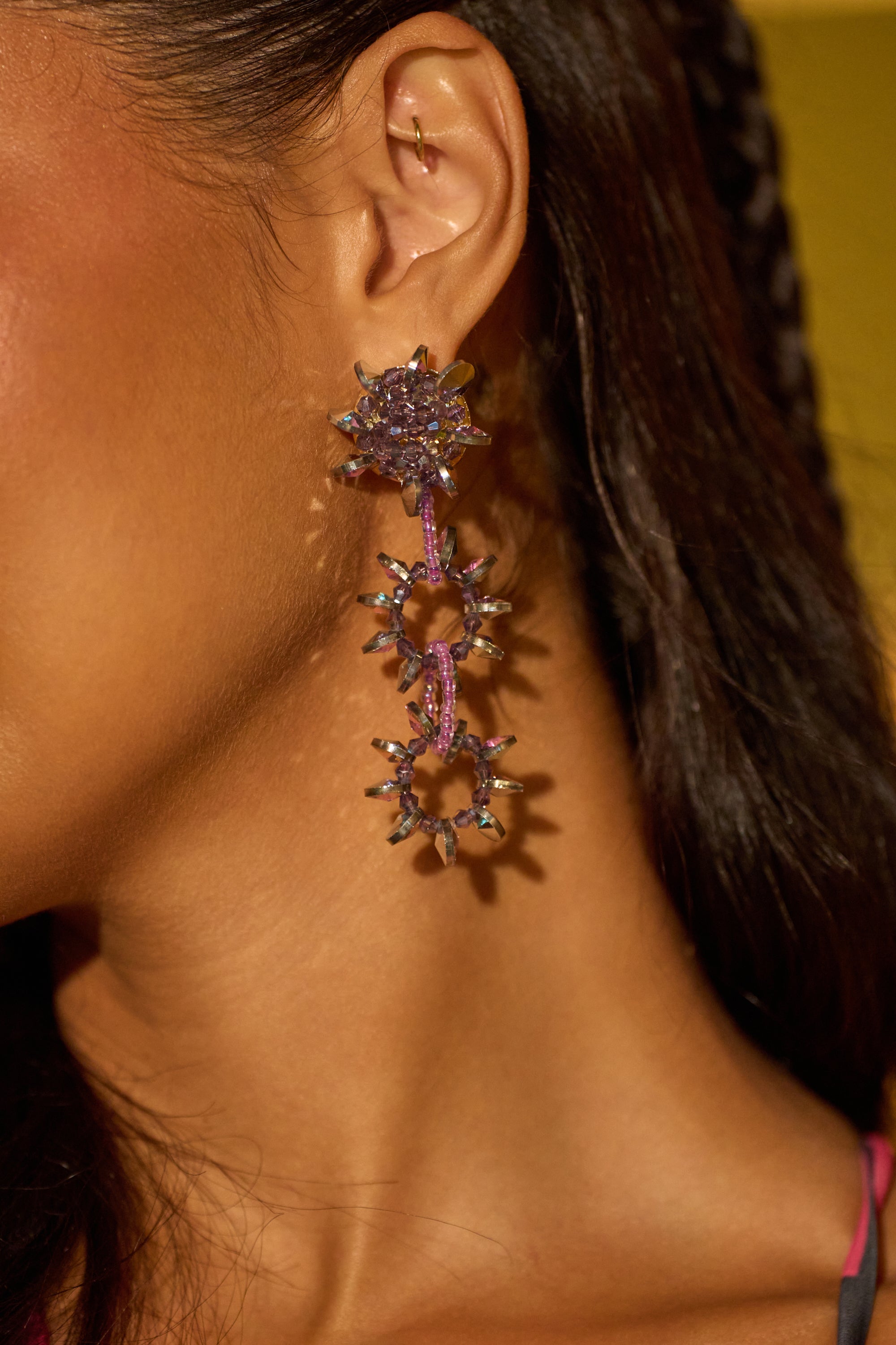 Close-up of a person’s ear wearing the TOO TRENDY EARRING—gold and purple floral drops with gemstone accents—ideal festival accessories, shown against a blurred yellow background.