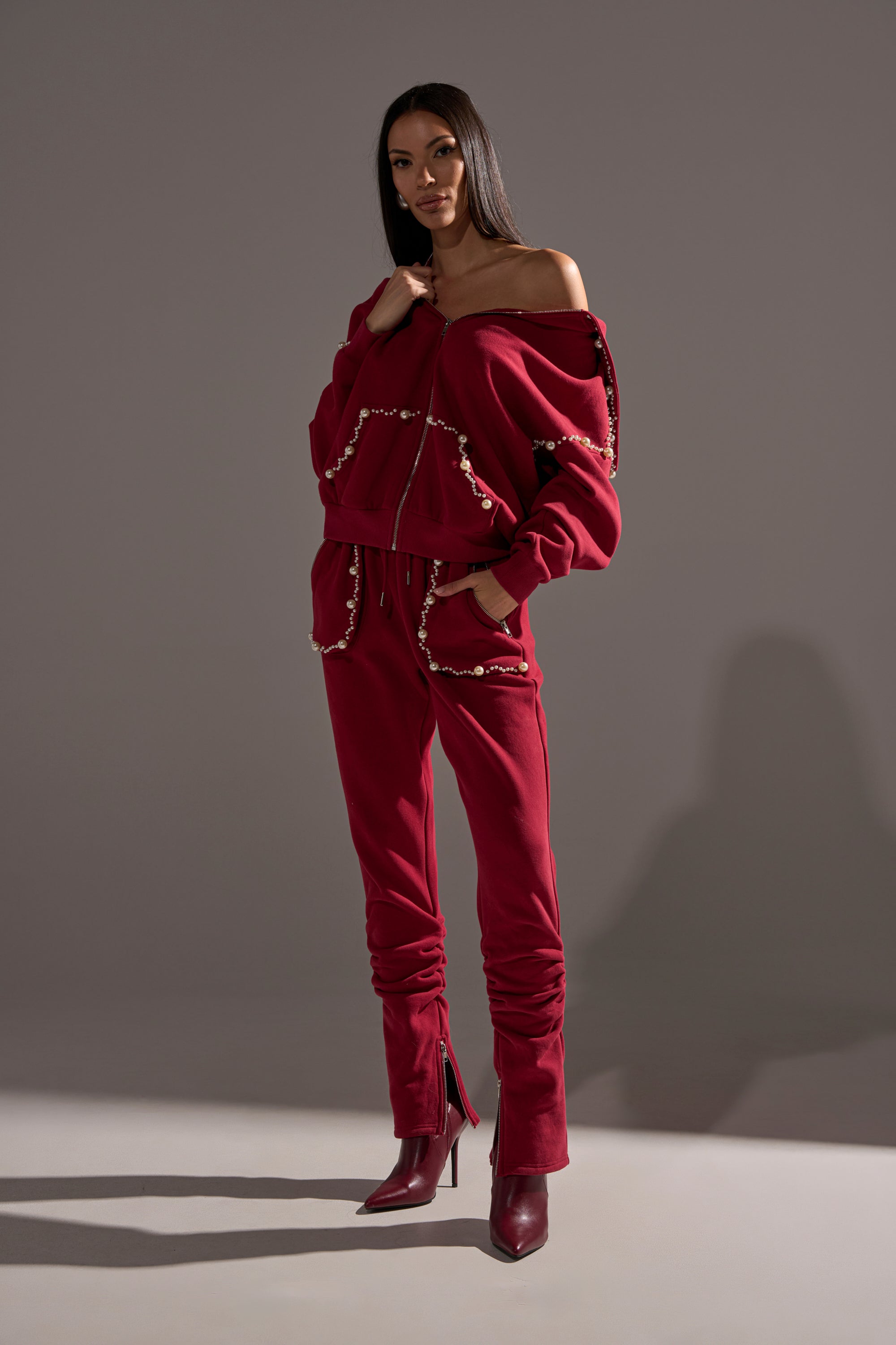 A woman models the COZY HOLIDAY PEARL JOGGER IN BURGUNDY—a stylish tracksuit with pearl accents, off-the-shoulder jacket, and matching pants—paired with maroon heeled boots against a plain gray background.