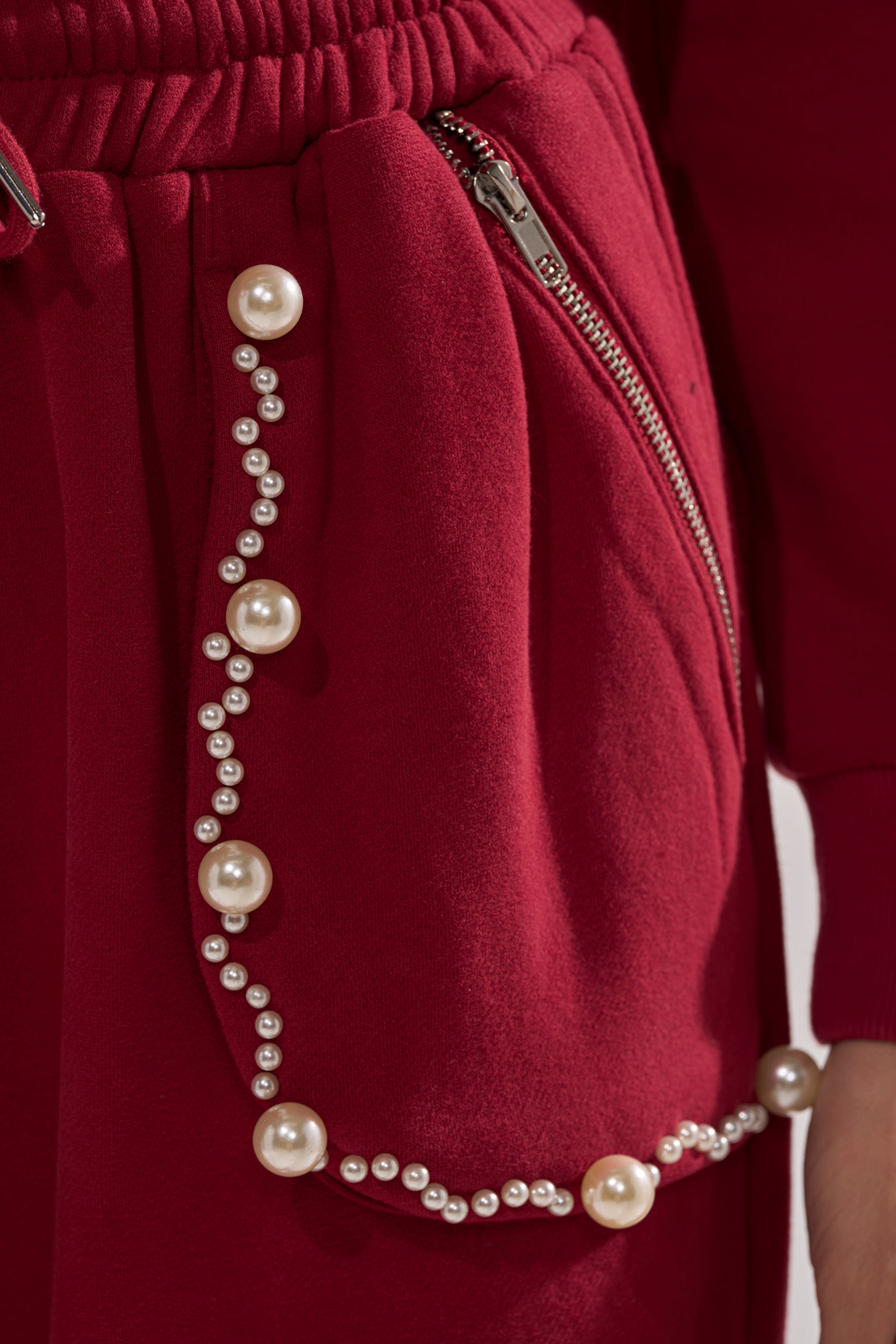Close-up of the COZY HOLIDAY PEARL JOGGER IN BURGUNDY, showcasing a visible zipper, decorative seam trim, and a mix of small and large pearl embellishments.