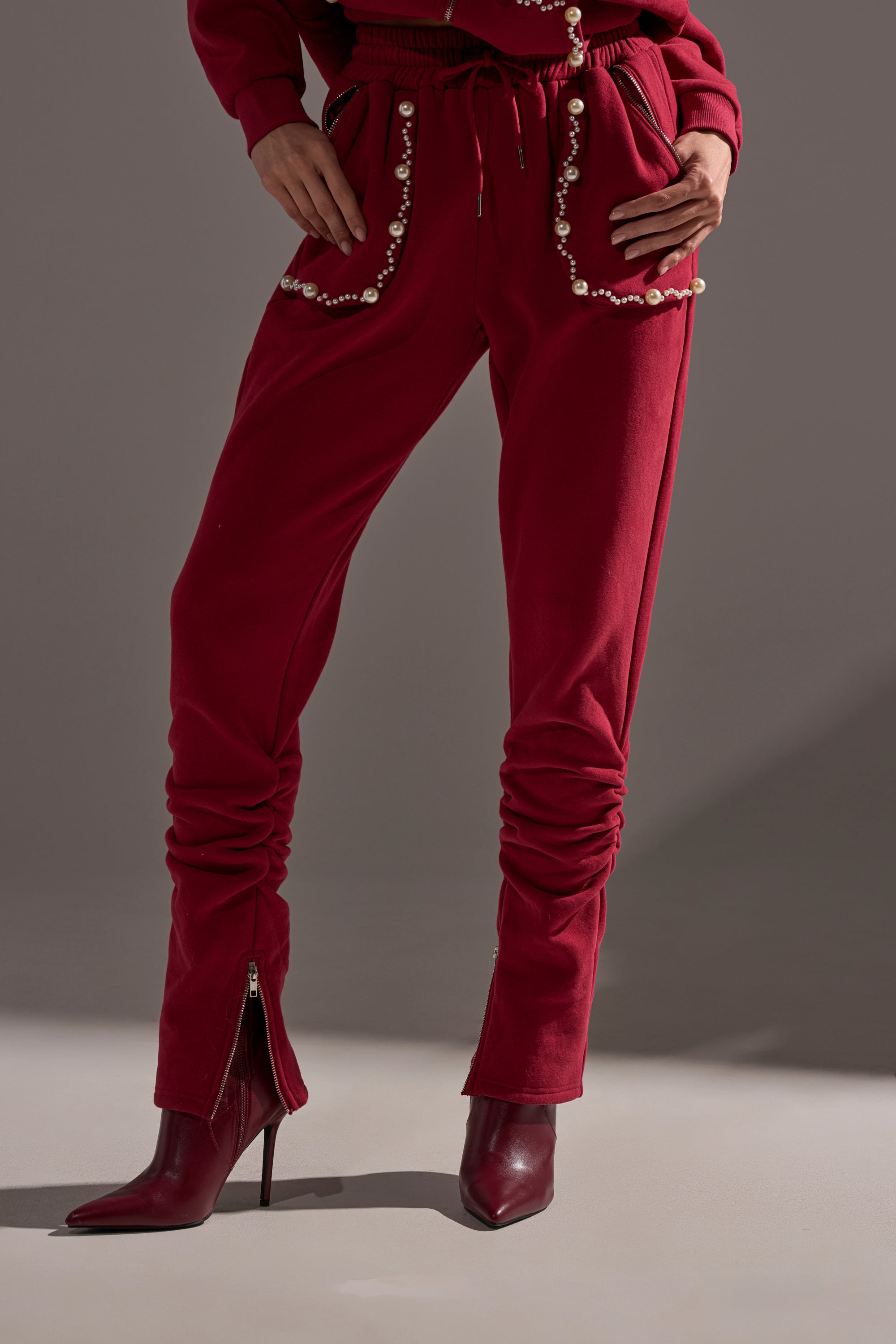 A person wearing the COZY HOLIDAY PEARL JOGGER IN BURGUNDY with pearl-embellished pockets and red heeled boots, shown from waist to feet, stands against a plain gray background.
