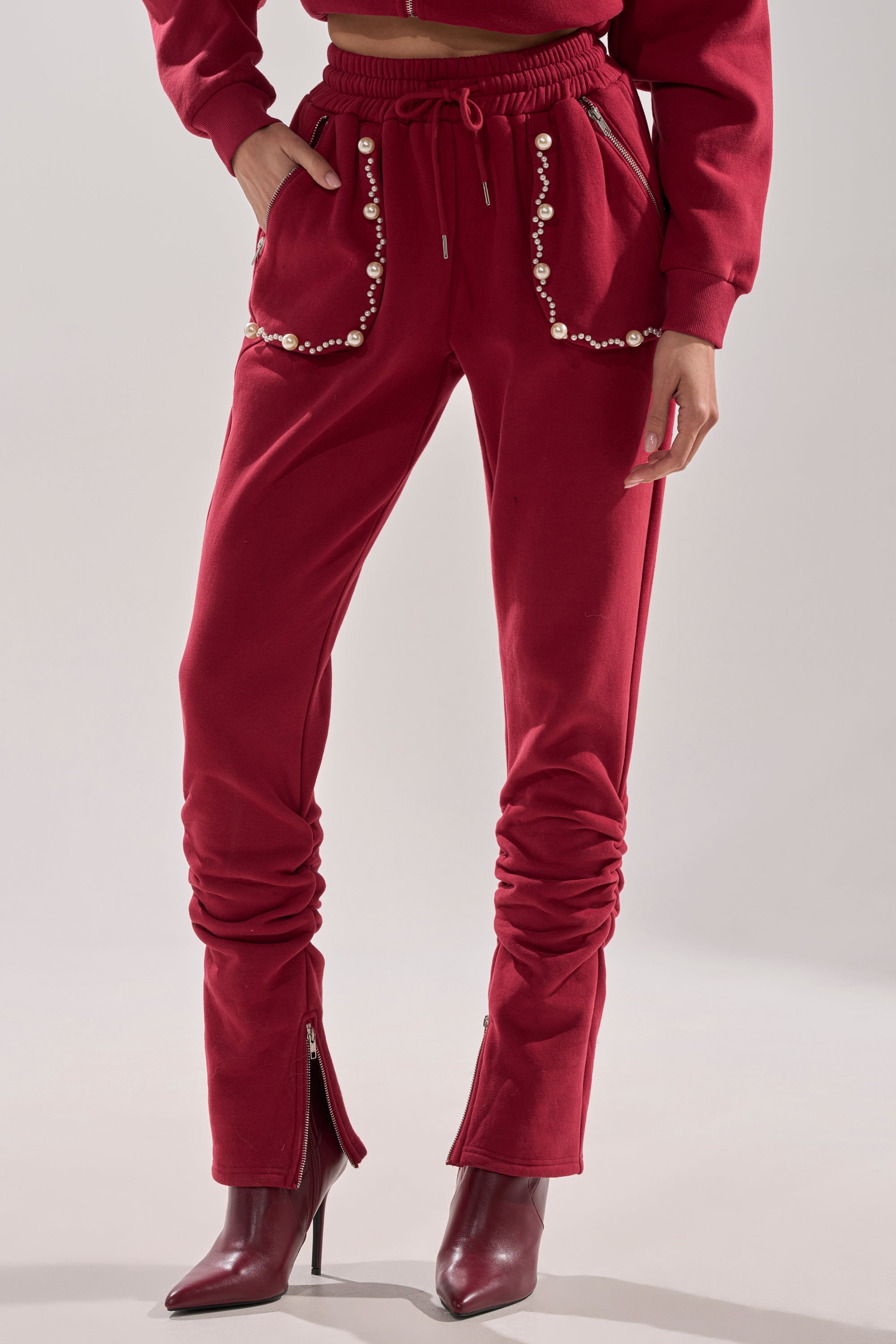 A person in the COZY HOLIDAY PEARL JOGGER IN BURGUNDY, featuring pearl-embellished pockets, ruching at the ankles, and zippered cuffs, styled with matching heeled boots and a glimpse of a red hoodie for a festive look.