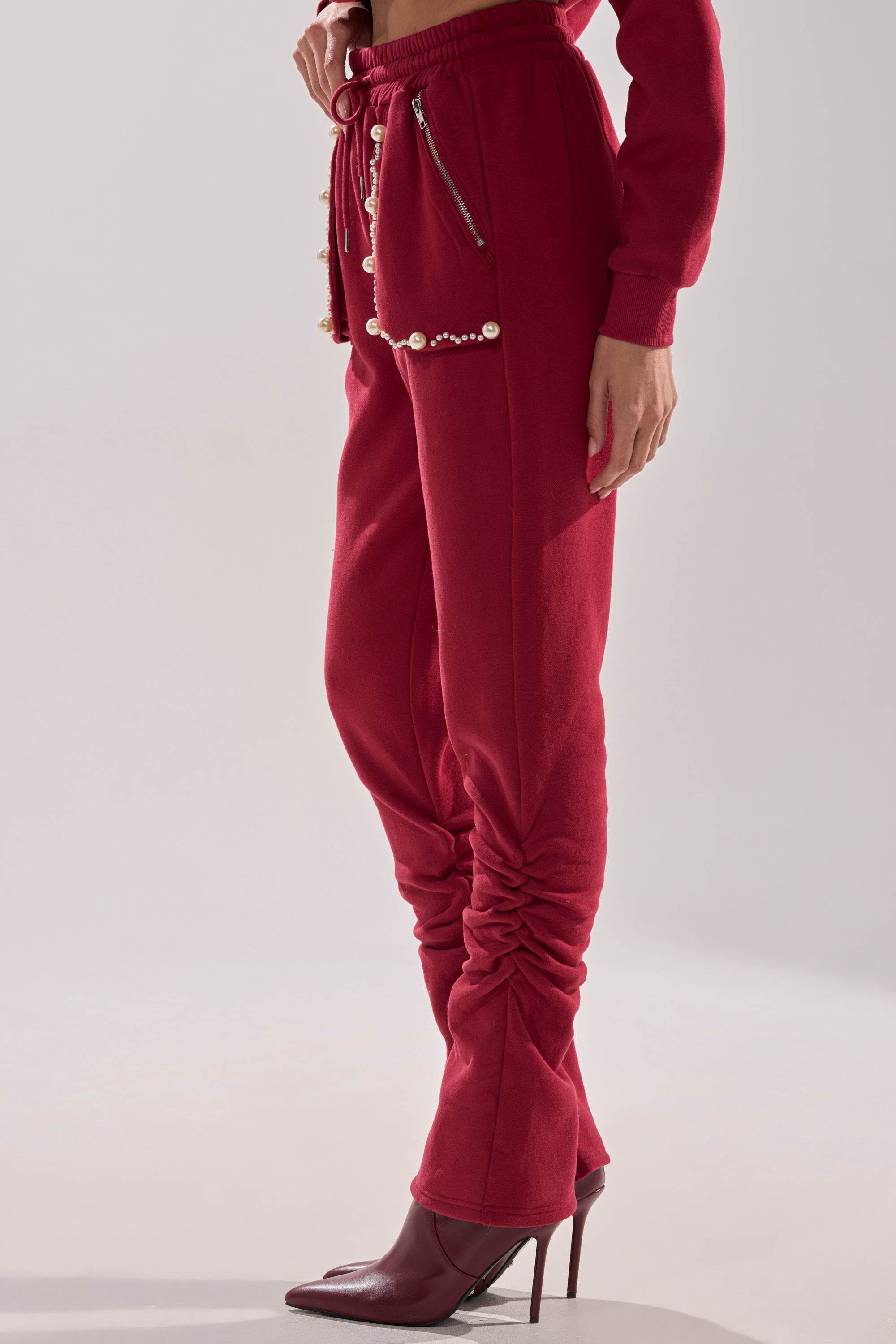 A person wears the COZY HOLIDAY PEARL JOGGER IN BURGUNDY with ruched legs, pearl and zipper waist details, and matching red heeled boots, standing against a plain light background.
