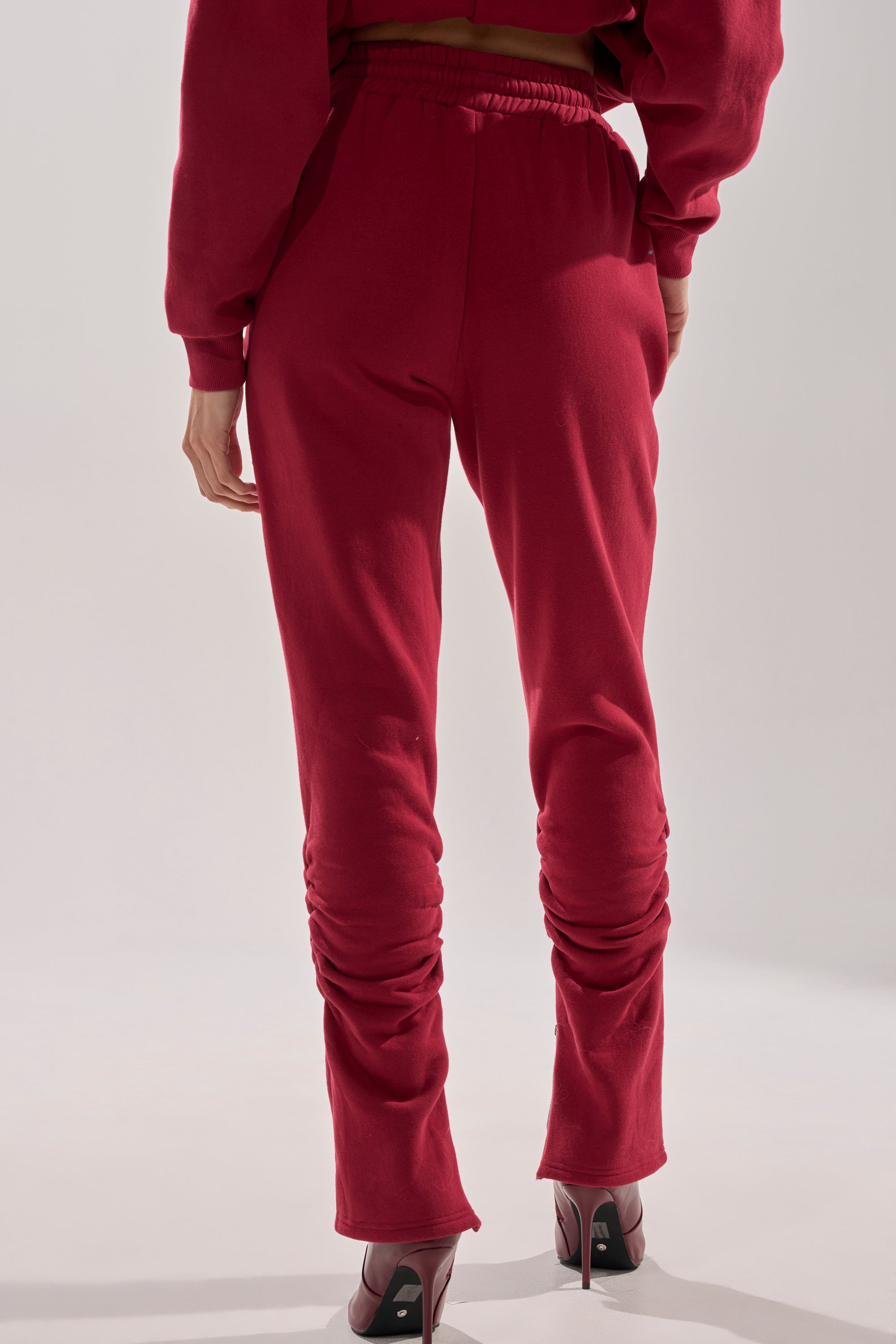 A person stands facing away from the camera in the COZY HOLIDAY PEARL JOGGER IN BURGUNDY and a matching red sweatshirt with high-heeled, open-toe shoes, set against a plain, light background.