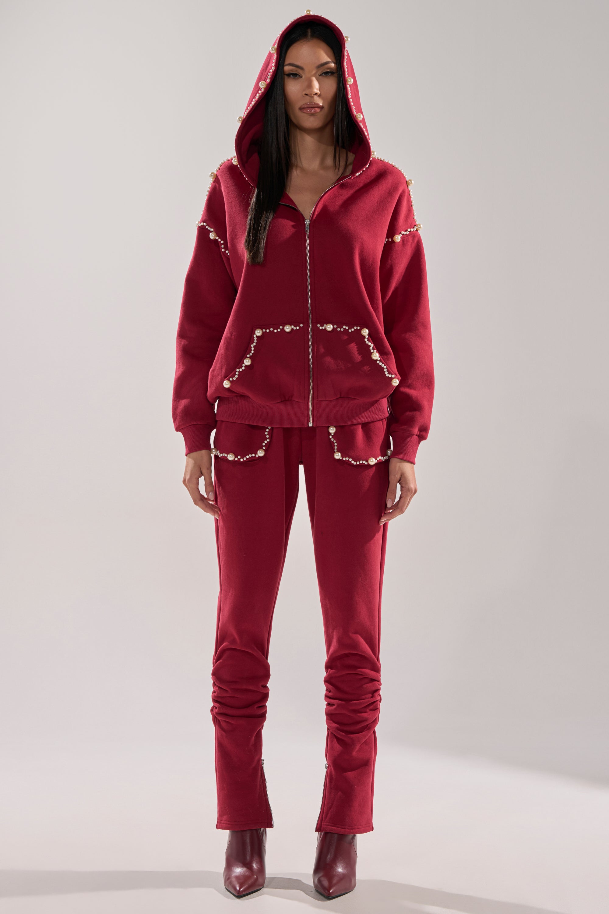A woman stands facing forward in the COZY HOLIDAY PEARL JOGGER IN BURGUNDY—matching deep red hoodie and joggers with pearl-like details on the shoulders, pockets, and seams—paired with burgundy heeled boots.