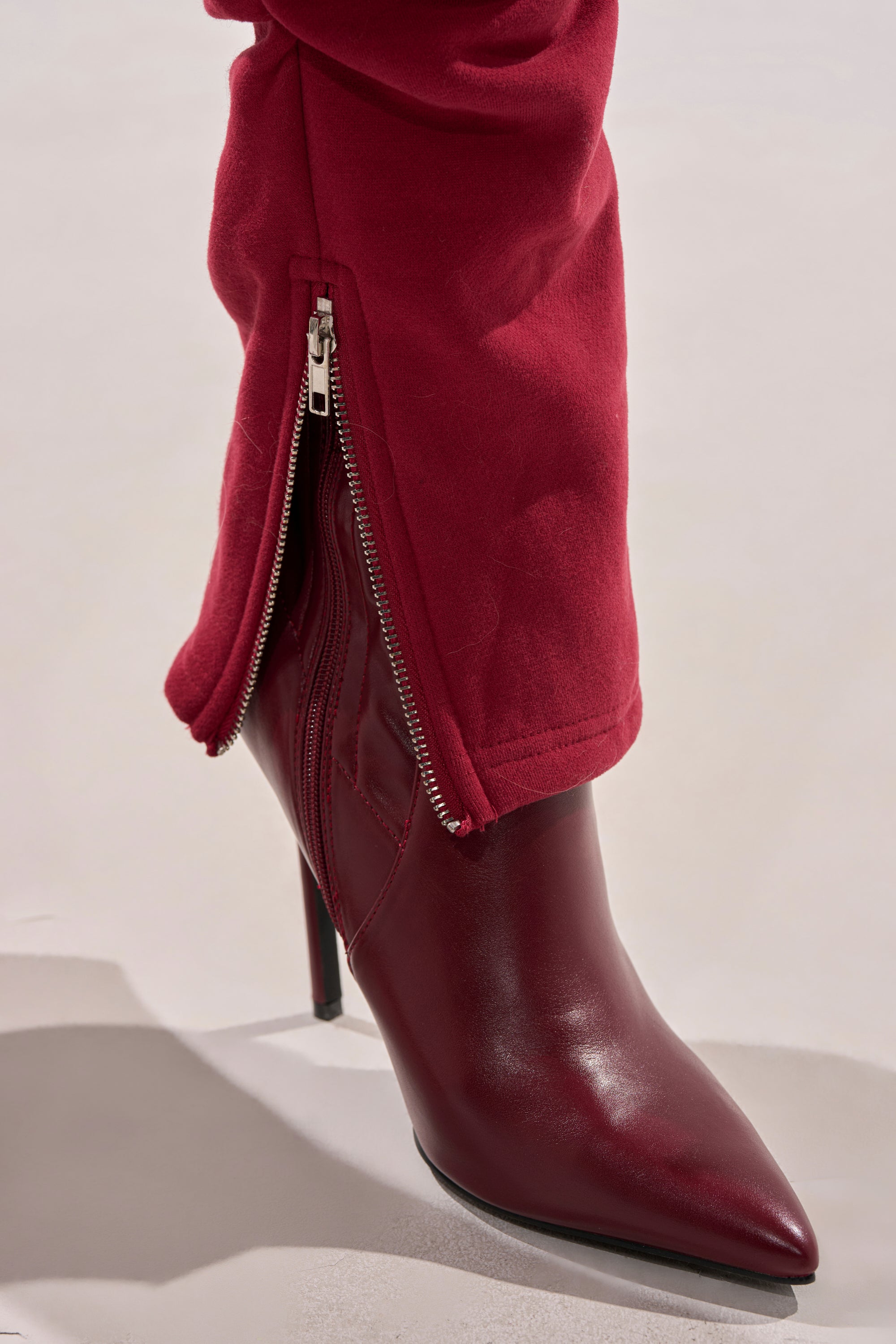 A close-up shows someone wearing the COZY HOLIDAY PEARL JOGGER IN BURGUNDY with a silver zipper partially open, revealing a red pointed-toe high-heeled boot against a minimal, neutral background.