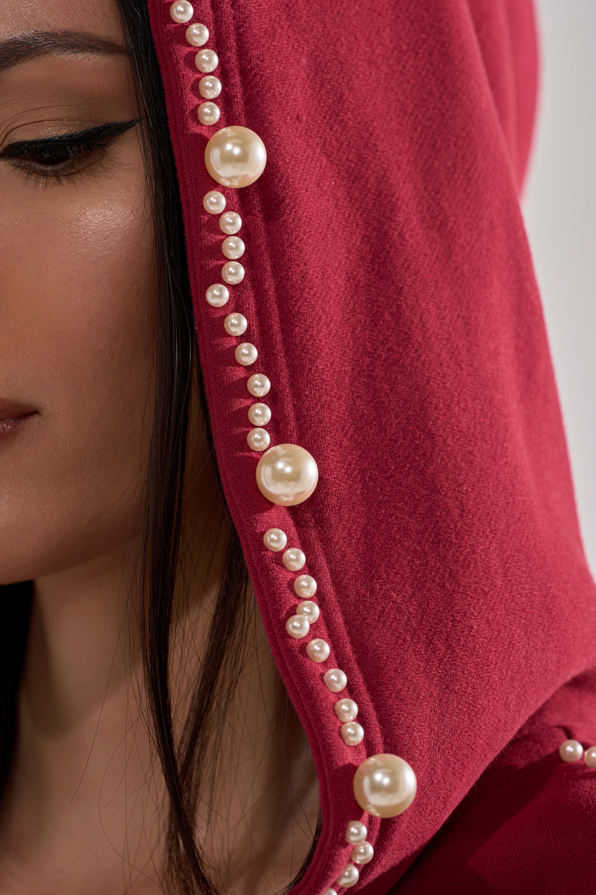 A woman wears the COZY HOLIDAY PEARL SWEATSHIRT IN BURGUNDY, featuring an oversized hood decorated with pearl-like beads. Only half her face with straight dark hair and subtle makeup is visible.