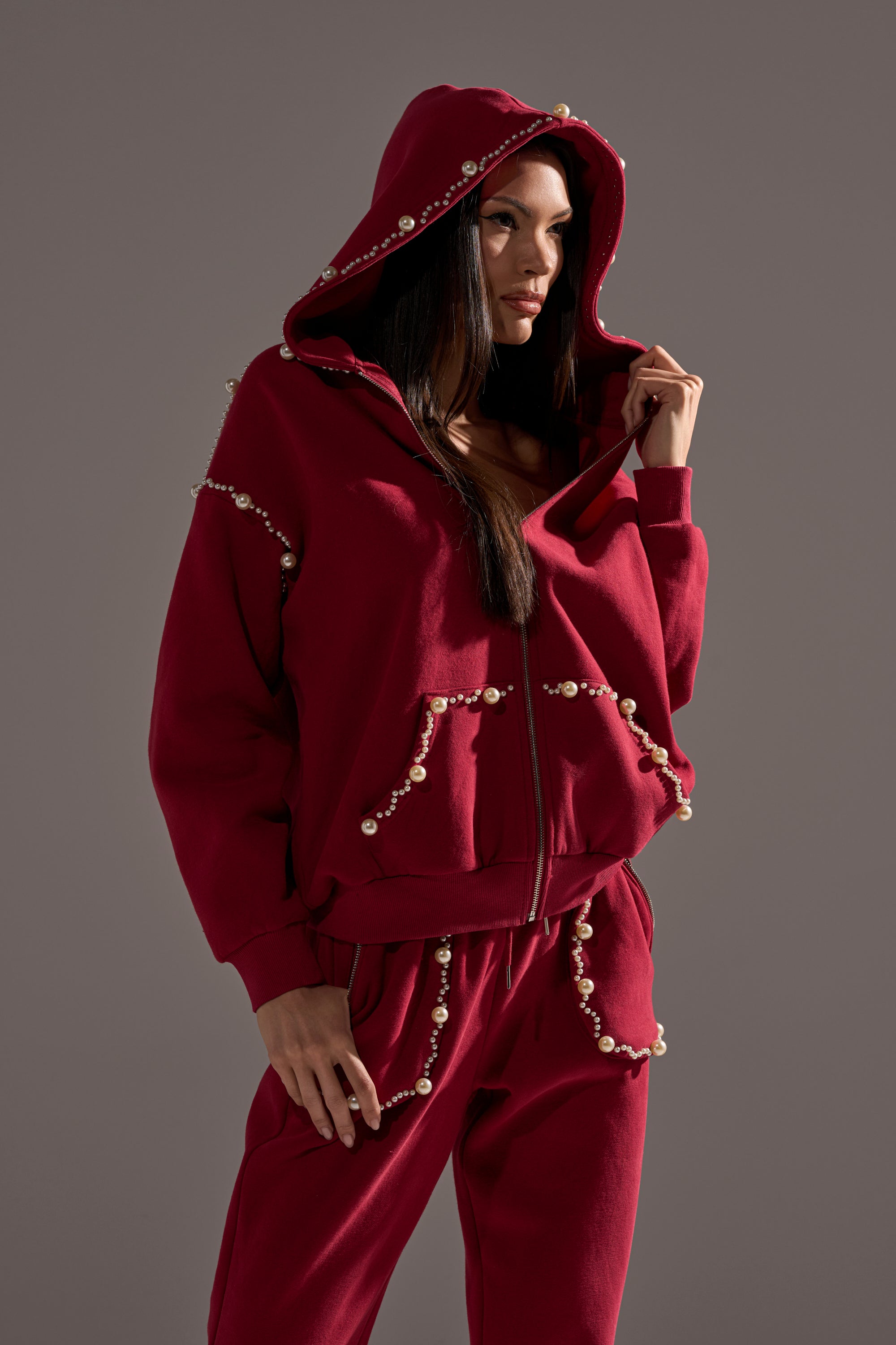 A woman wearing the COZY HOLIDAY PEARL SWEATSHIRT IN BURGUNDY, adorned with pearls, stands against a plain gray background and looks slightly to the side.