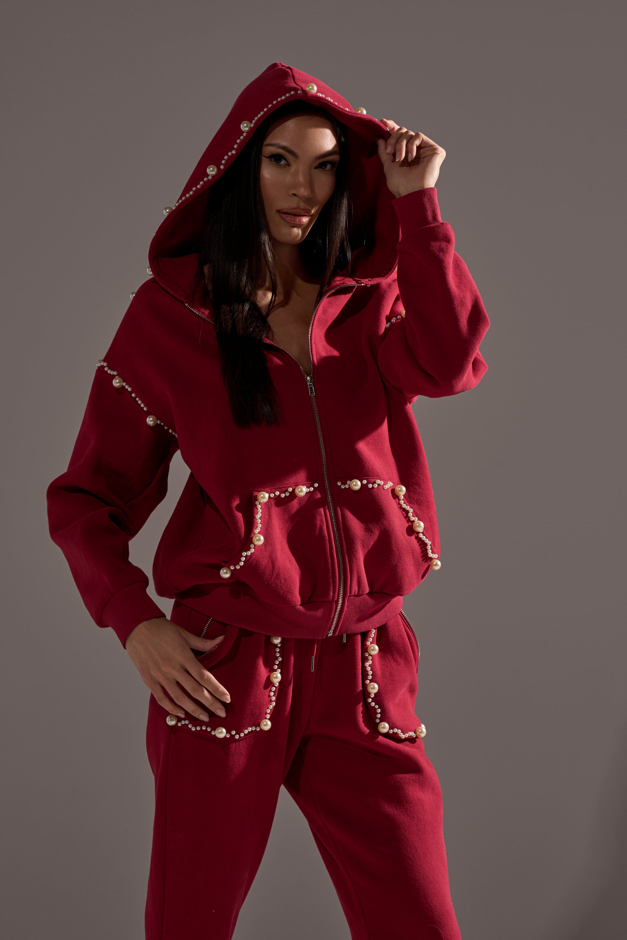 A woman in the COZY HOLIDAY PEARL SWEATSHIRT IN BURGUNDY poses with one hand holding her hood, set against a plain gray background.