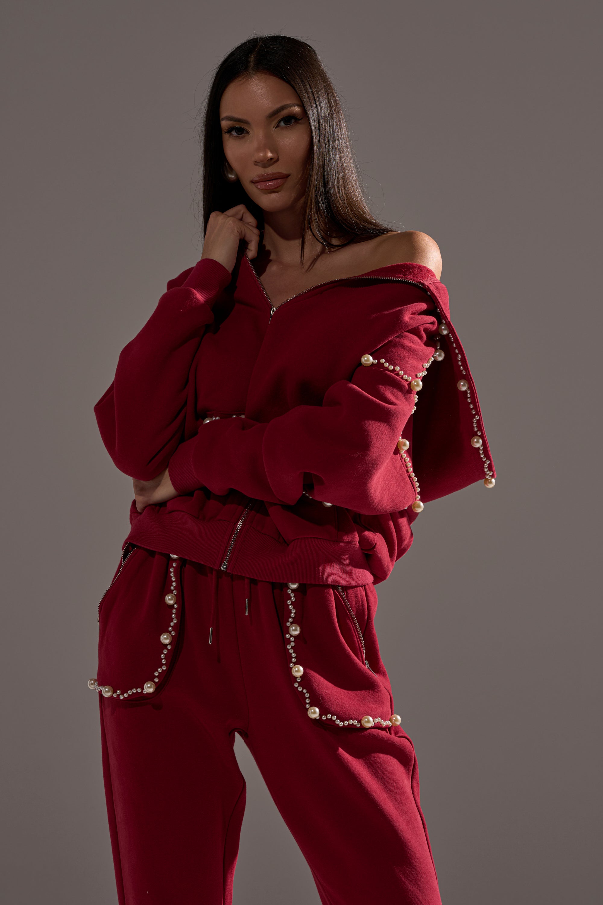 A woman models the COZY HOLIDAY PEARL SWEATSHIRT IN BURGUNDY, a stylish red top adorned with pearls on the sleeves and pockets, as she poses confidently against a plain gray background.