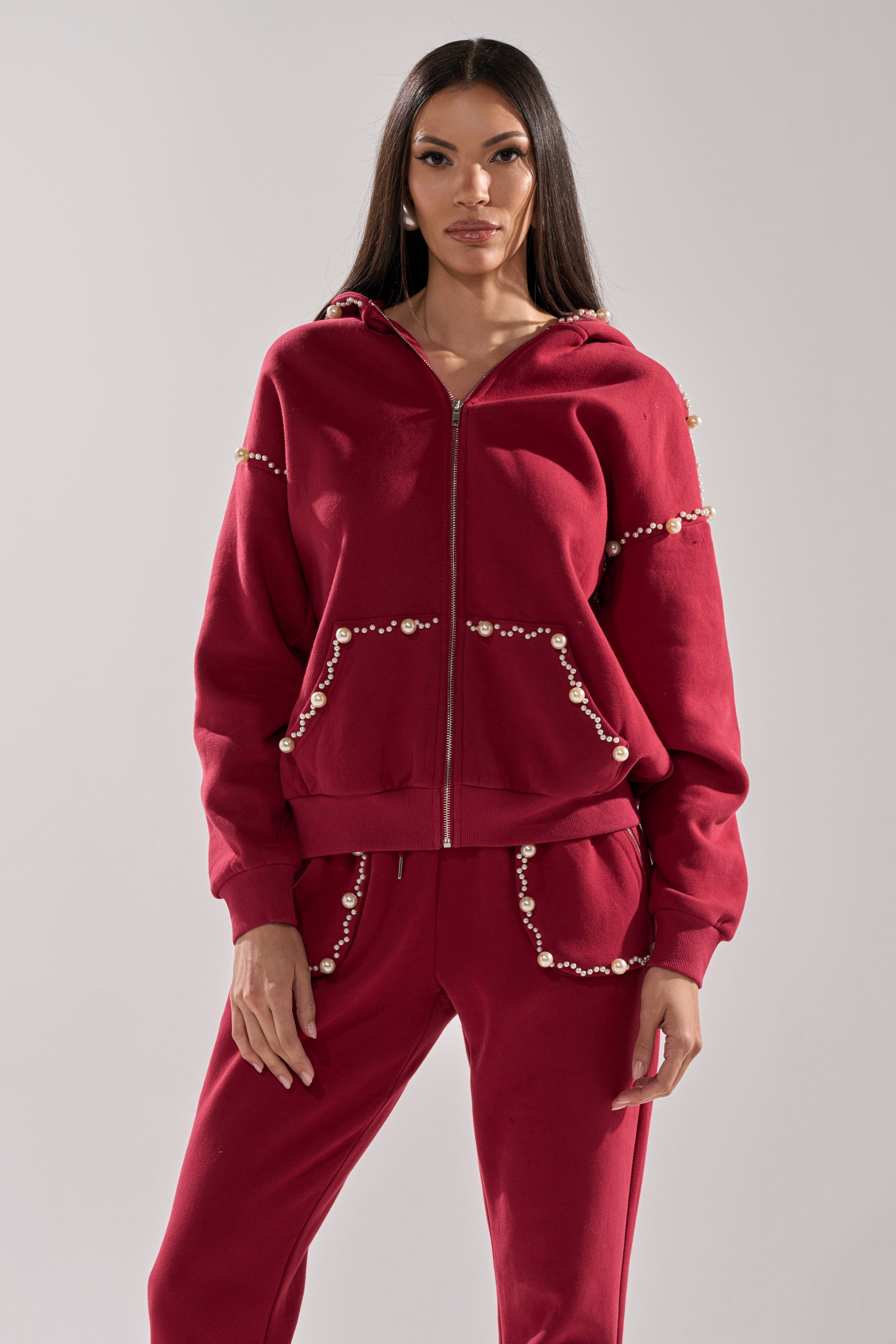 A woman faces the camera in a COZY HOLIDAY PEARL SWEATSHIRT IN BURGUNDY, featuring pearl-like beads along the seams and pockets, paired with matching sweatpants against a plain light background.