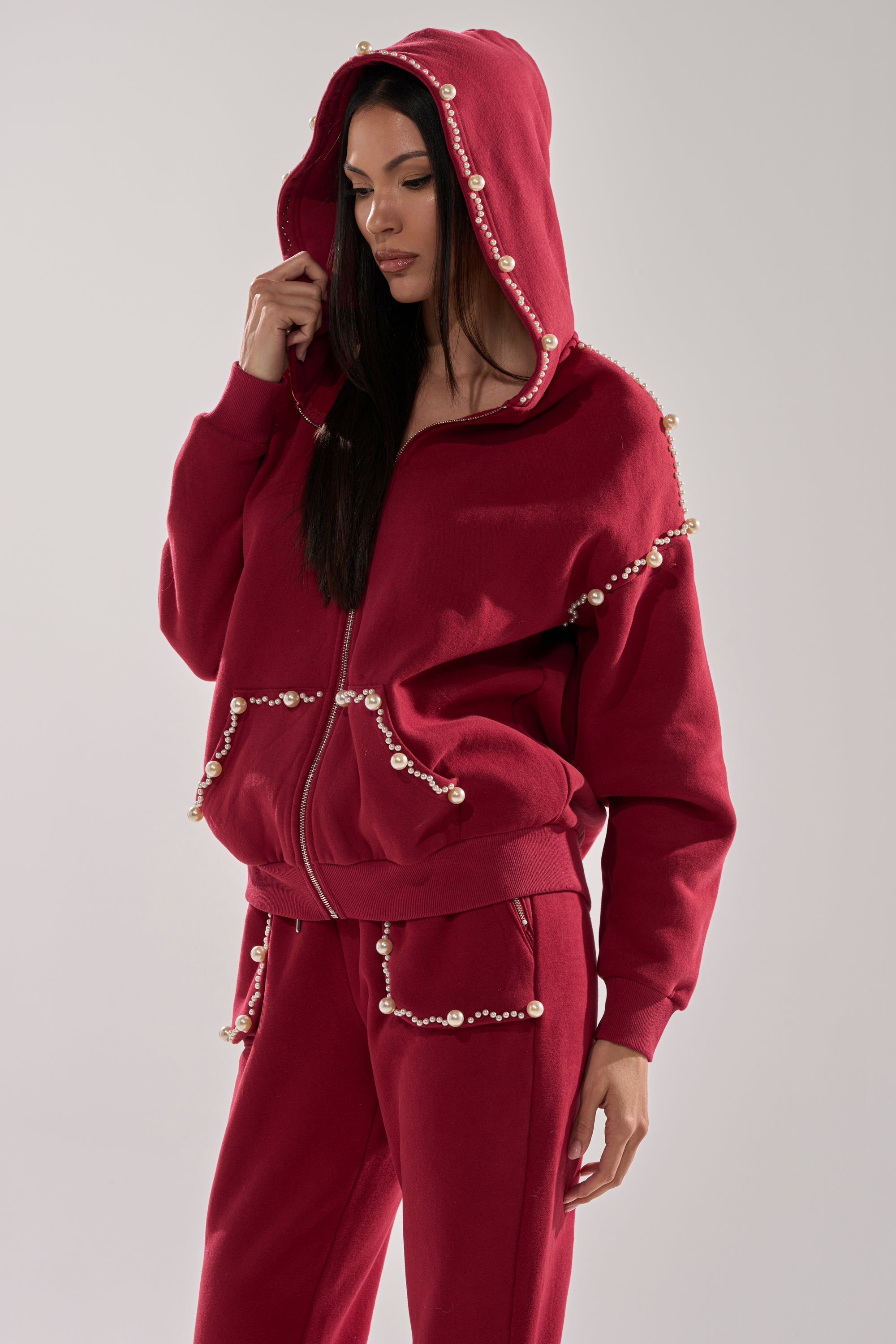 A woman stands against a plain background, wearing the COZY HOLIDAY PEARL SWEATSHIRT IN BURGUNDY with matching pants. Both pieces feature pearl-like bead accents along the edges as she gently touches the hood, showcasing the festive, cozy style.