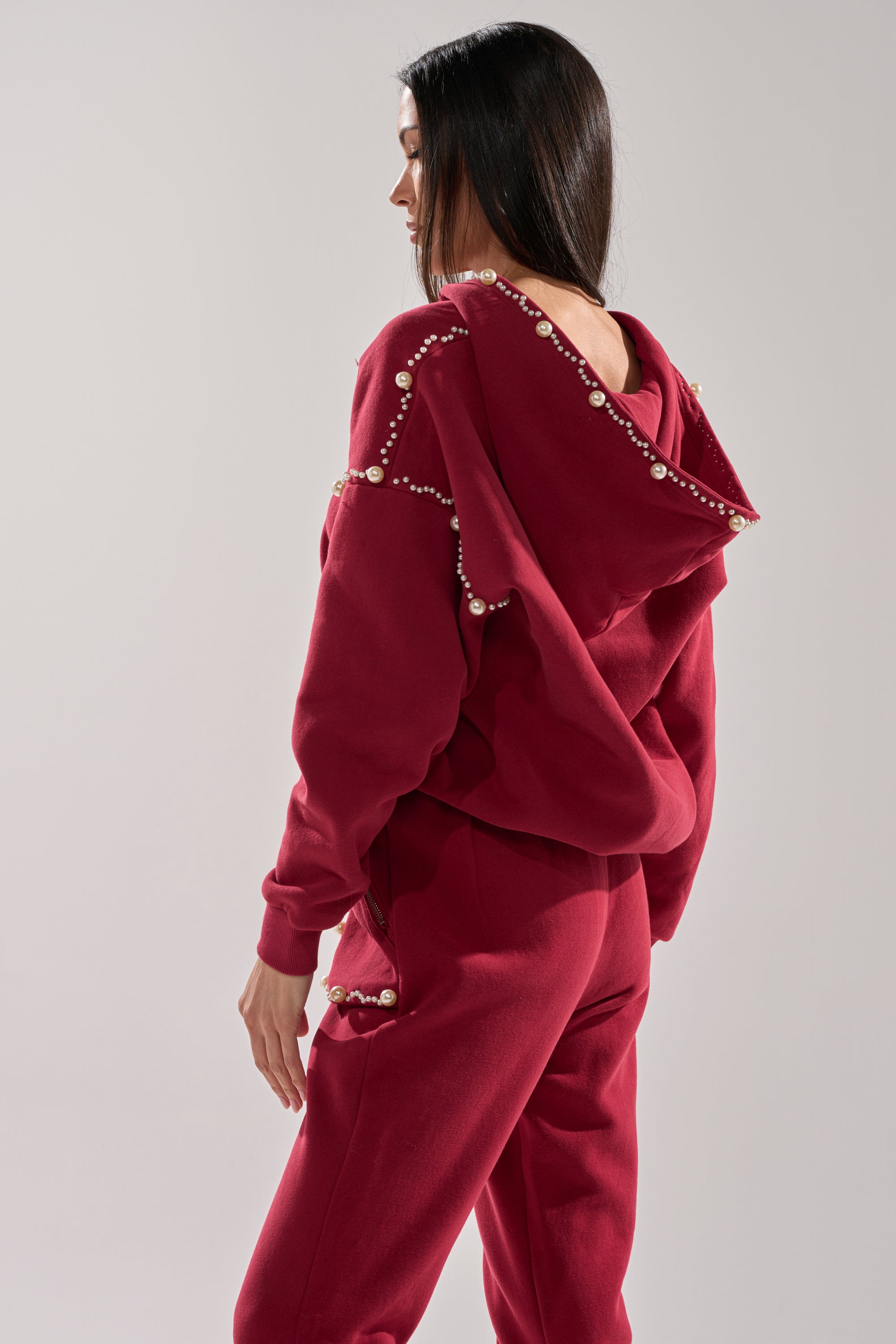 A woman stands sideways against a light gray background wearing the COZY HOLIDAY PEARL SWEATSHIRT IN BURGUNDY, a red zip-up tracksuit decorated with pearls along the hood, shoulders, and sleeves.
