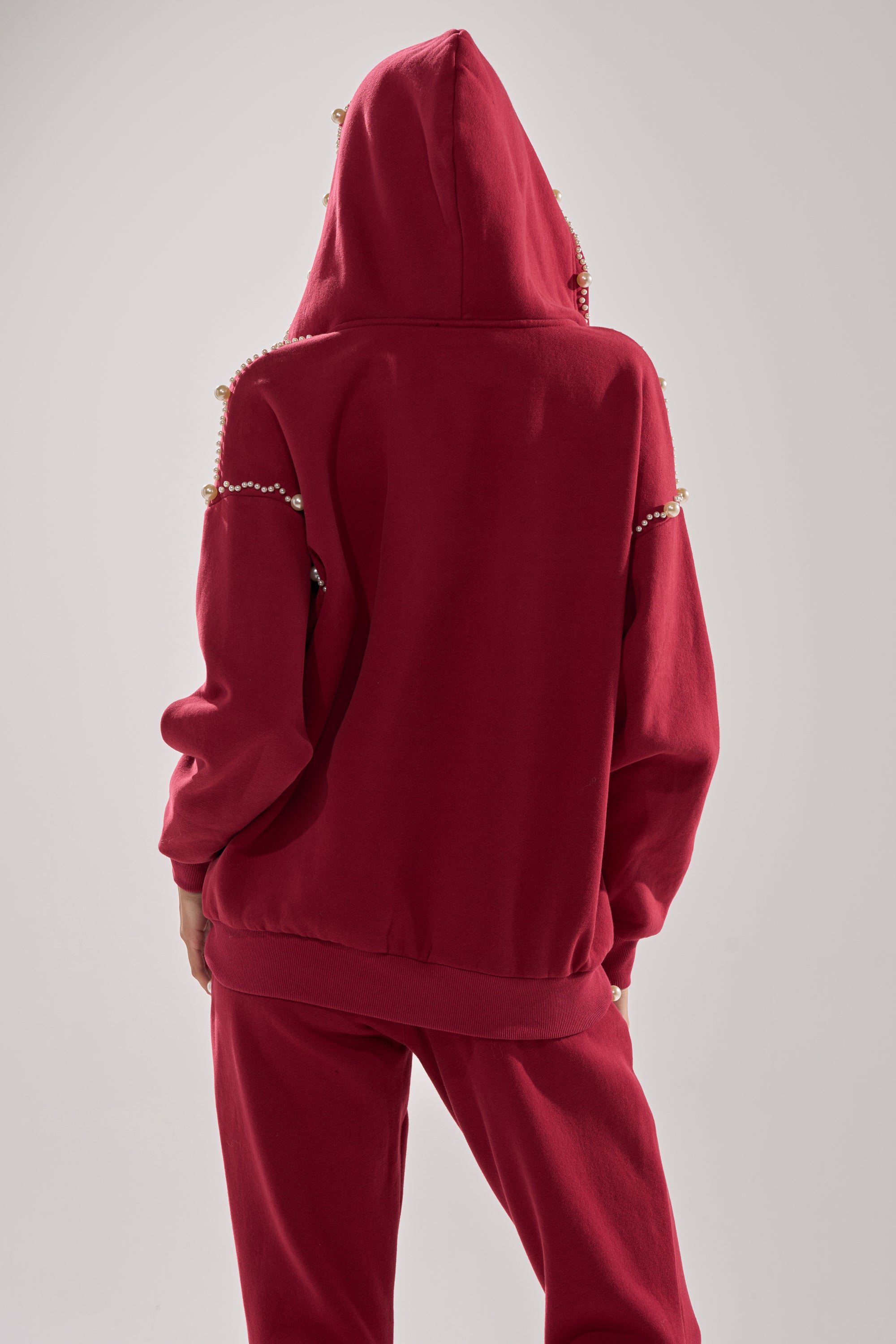 A person faces away from the camera against a plain light backdrop, wearing the COZY HOLIDAY PEARL SWEATSHIRT IN BURGUNDY—an oversized, festive zip-up with pearl embellishments—paired with matching red pants.