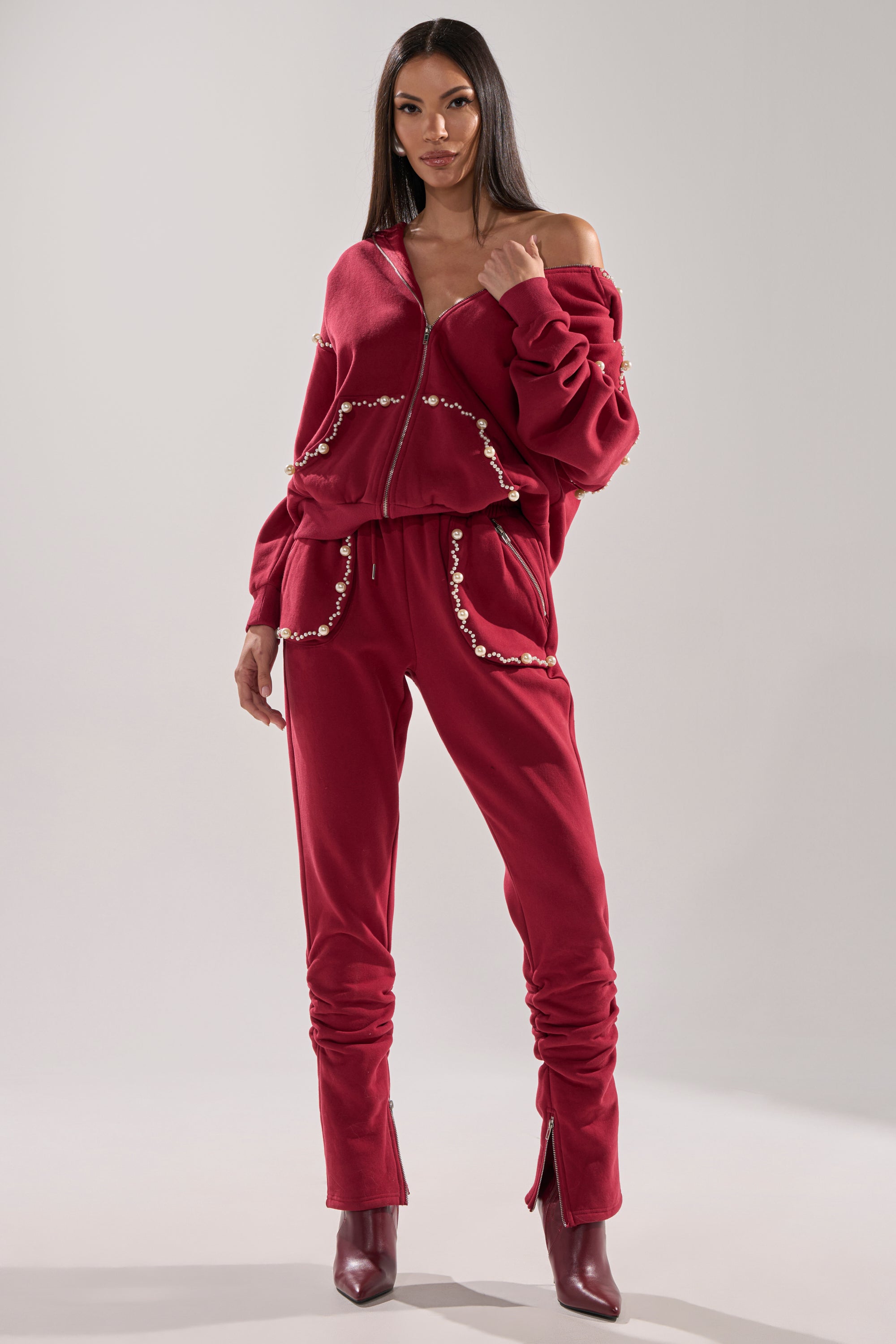 A woman stands confidently in the COZY HOLIDAY PEARL SWEATSHIRT IN BURGUNDY, paired with matching pants and dark red ankle boots, slightly off one shoulder, posing against a plain light gray background.