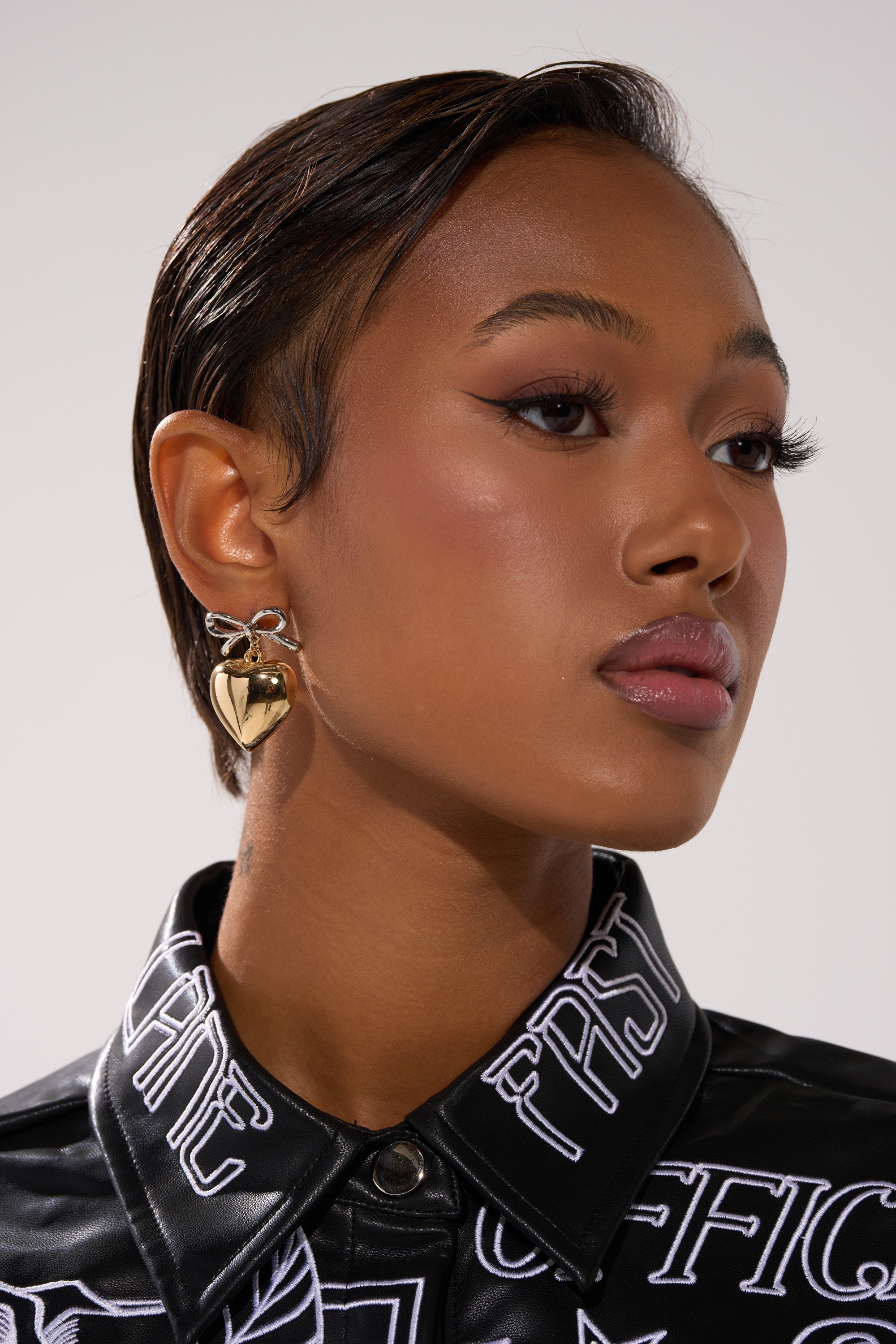 A woman with sleek, short hair and winged eyeliner models the BIG HEART EARRING—bold gold hearts—paired with a black button-up shirt featuring white graphic text, gazing confidently to the side against a plain background. 1
