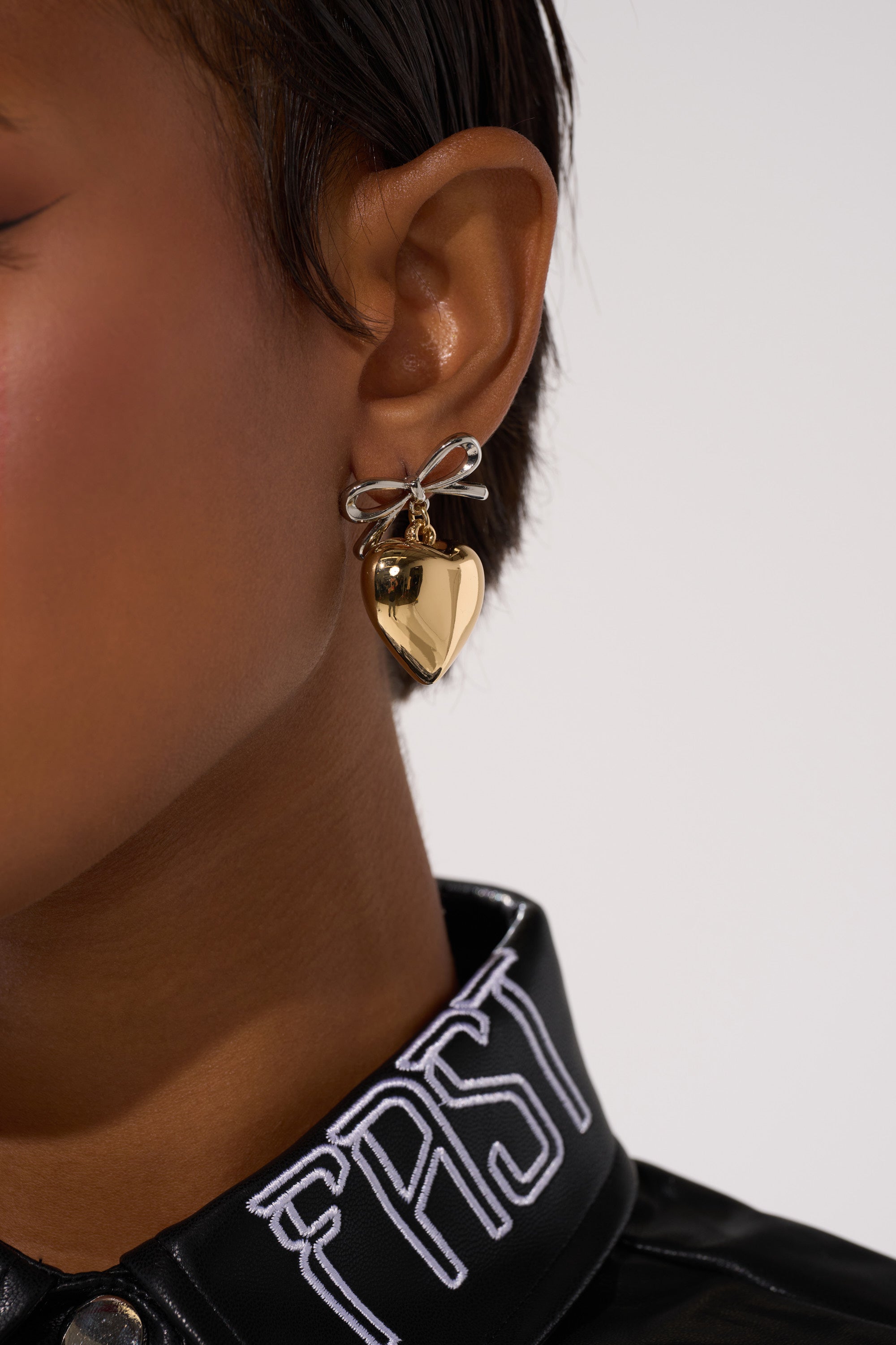 Close-up of a person with short hair wearing the BIG HEART EARRING—shiny gold hearts with silver bows. They're dressed in a black jacket featuring white "FAST" embroidery on the collar. 3