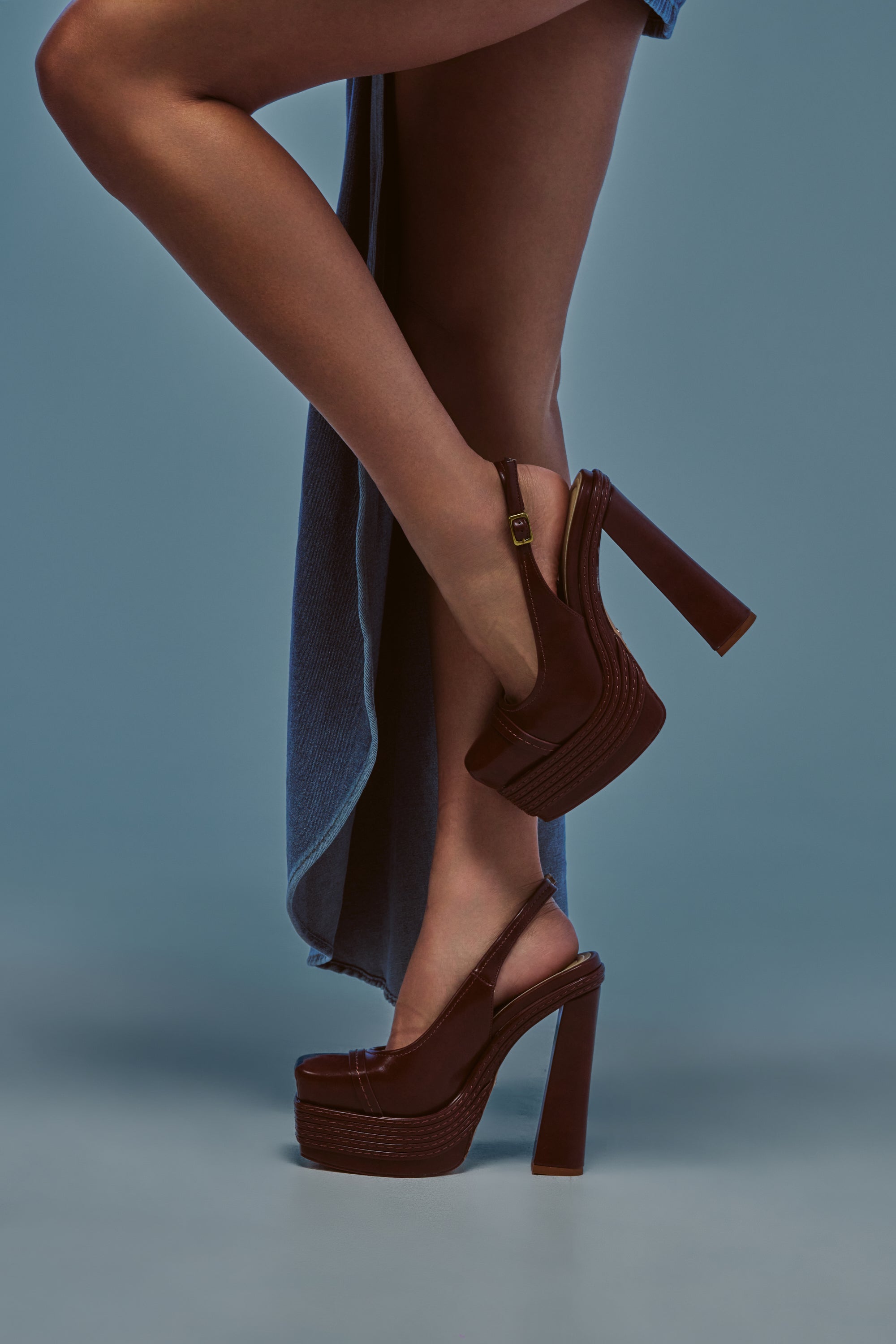 Wearing the AZALEA WANG PEGGY BROWN PU BLOCK PUMP—a stylish brown leather platform with a chic slingback strap—a person poses in a blue skirt, standing with one leg crossed over the other against a blue background.
