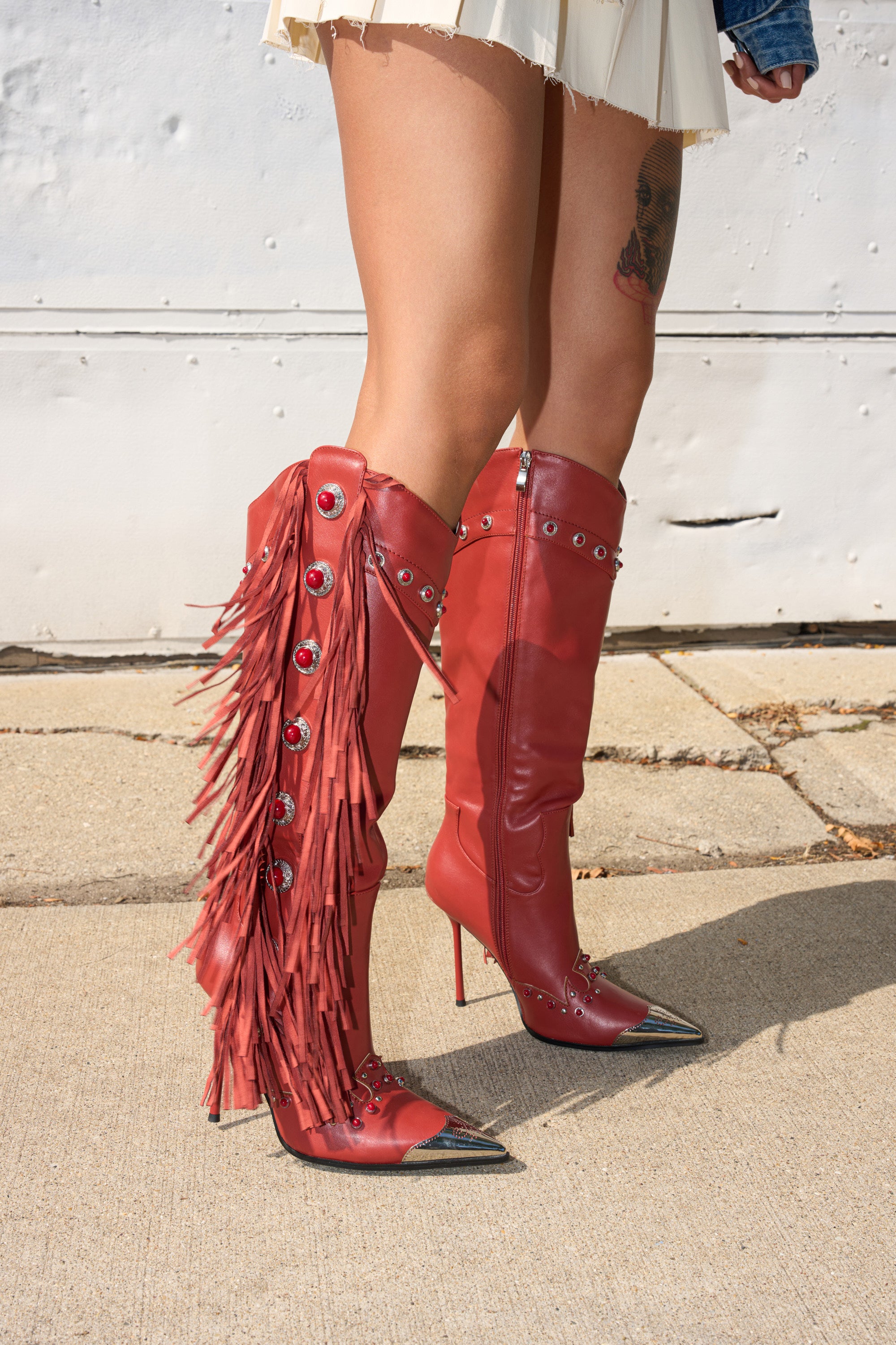 A person in a cream pleated skirt stands on a sidewalk wearing the AZALEA WANG PELAGIANA Red Western Pointed Toe Stiletto Boot, featuring fringe, metal studs, and knee-high style. 1