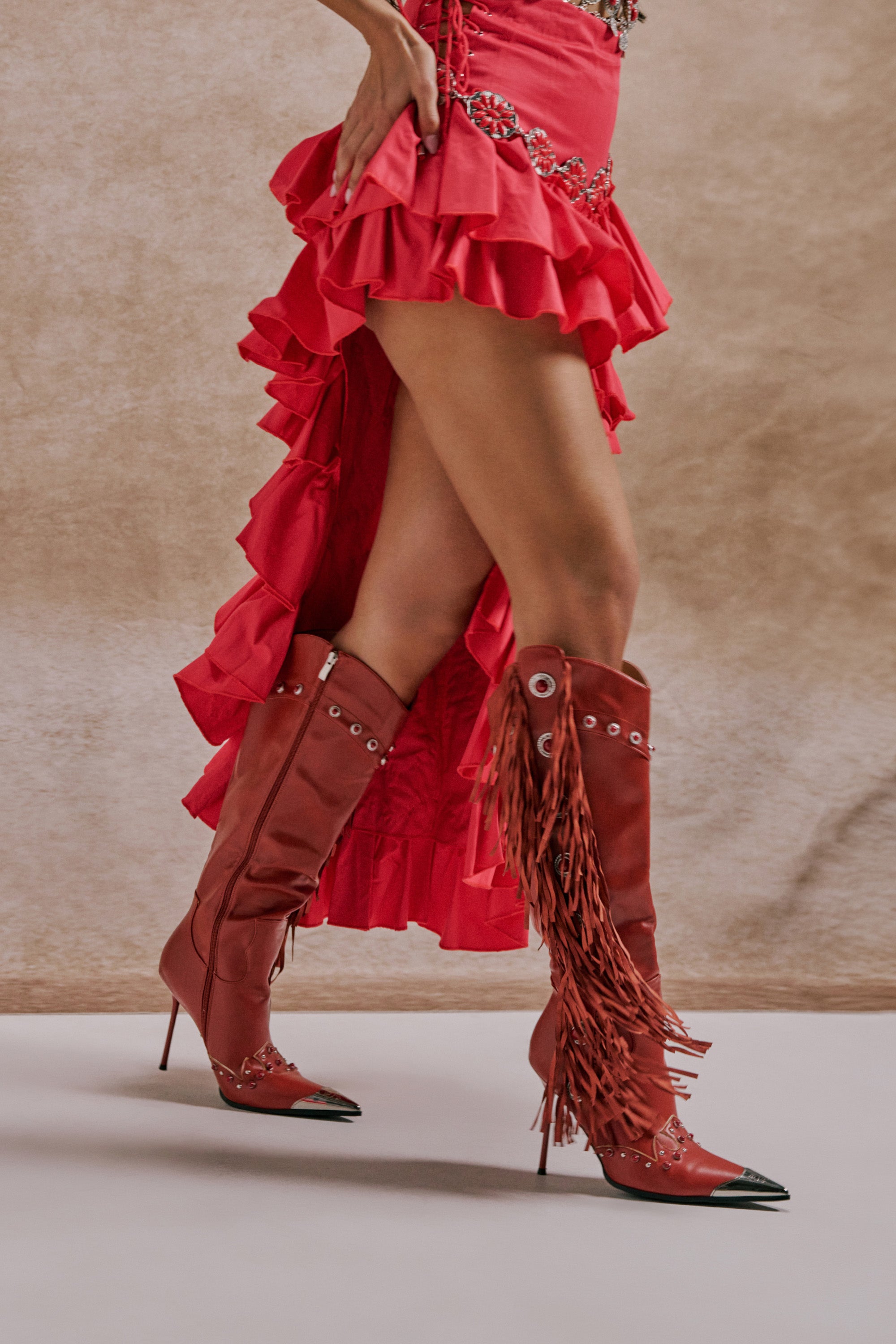 Wearing a ruffled high-low red skirt and AZALEA WANG PELAGIANA RED WESTERN POINTED TOE STILETTO BOOTS with fringe, a person stands against a neutral background. 0