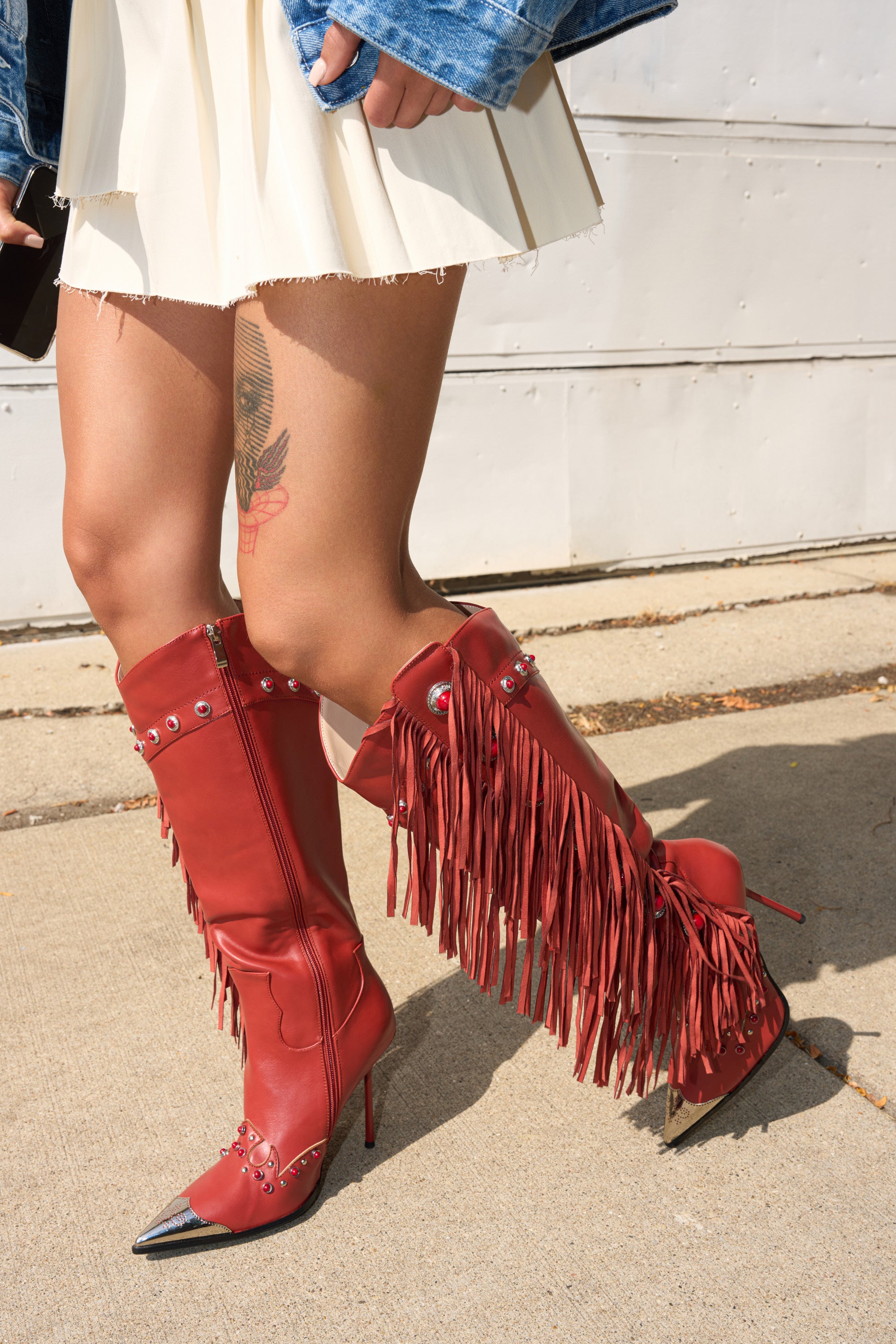 A person in a white skirt, blue denim jacket, and AZALEA WANG PELAGIANA RED WESTERN POINTED TOE STILETTO BOOTS with silver accents stands on the sidewalk. A tattoo is visible on their upper leg. 3