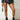 A person stands against a white brick wall, dressed in a studded denim jacket, a black pleated mini skirt, and the AZALEA WANG PEMA BLACK FLAT BOOTIE featuring silver accents. They carry a small silver chain bag, and the wooden floor is visible.