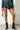 A person stands against a white brick wall, dressed in a studded denim jacket, a black pleated mini skirt, and the AZALEA WANG PEMA BLACK FLAT BOOTIE featuring silver accents. They carry a small silver chain bag, and the wooden floor is visible.