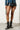 A person is standing against a white brick wall, wearing a studded denim jacket and a black pleated faux leather skirt with ring details. Their AZALEA WANG PEMA BLACK FLAT BOOTIE perfectly complements the edgy look as they hold a small chain purse.