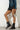 A person stands against a white brick wall, wearing a blue studded jacket and a black faux leather skirt. They have on the AZALEA WANG PEMA BLACK FLAT BOOTIE, which features lace-up combat styling with metallic toe caps. They carry a small silver beaded handbag, and their face is not visible.
