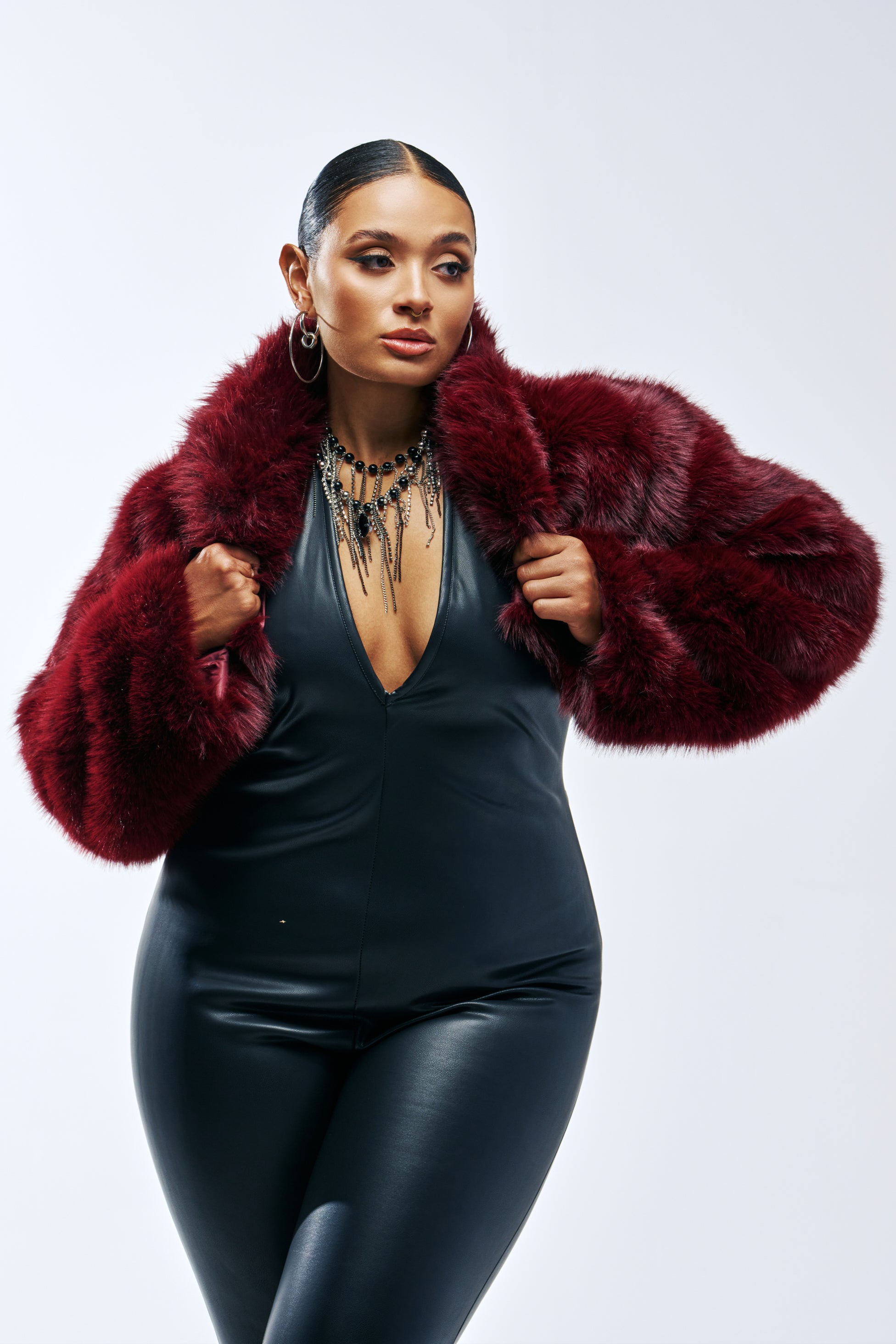 A woman wears a burgundy PENNY FAUX FUR SHRUG with a cropped silhouette over a black jumpsuit.