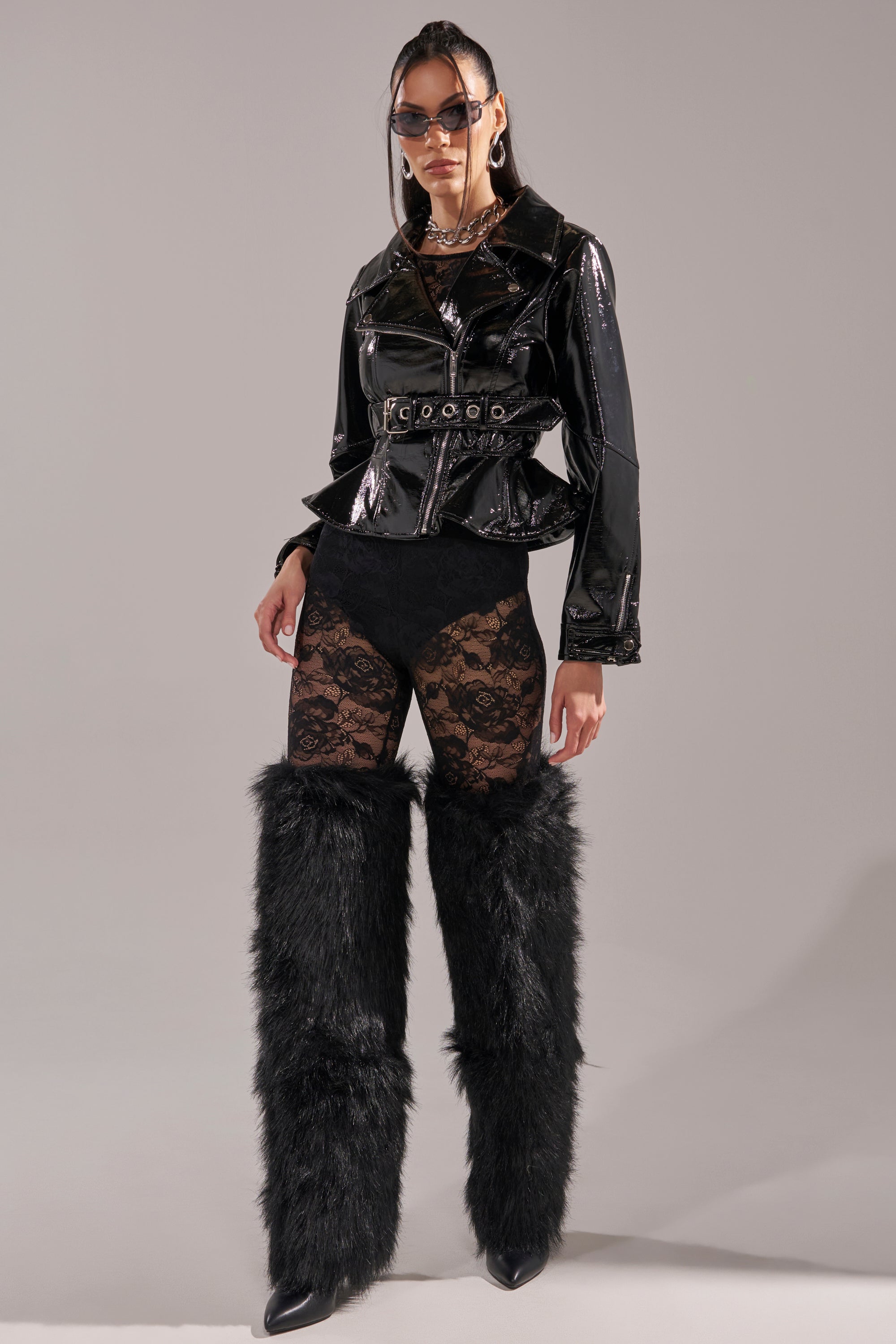 Wearing the PEP IN MY STEP PATENT MOTO jacket, black lace bodysuit, large black sunglasses, and furry leg warmers with pointed shoes, a person poses confidently against a plain background. 10
