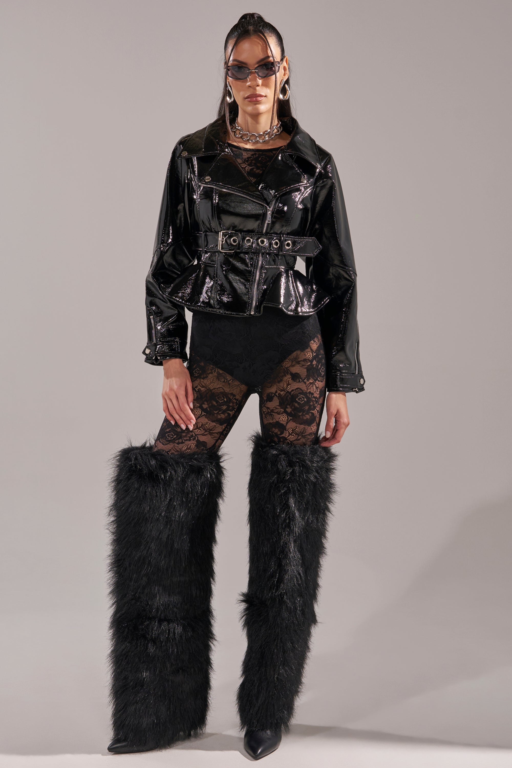 Wearing sunglasses, high-waisted bottoms, black lace tights, tall black furry leg warmers, and the PEP IN MY STEP PATENT MOTO jacket with a belt, a person stands against a plain gray background. 8