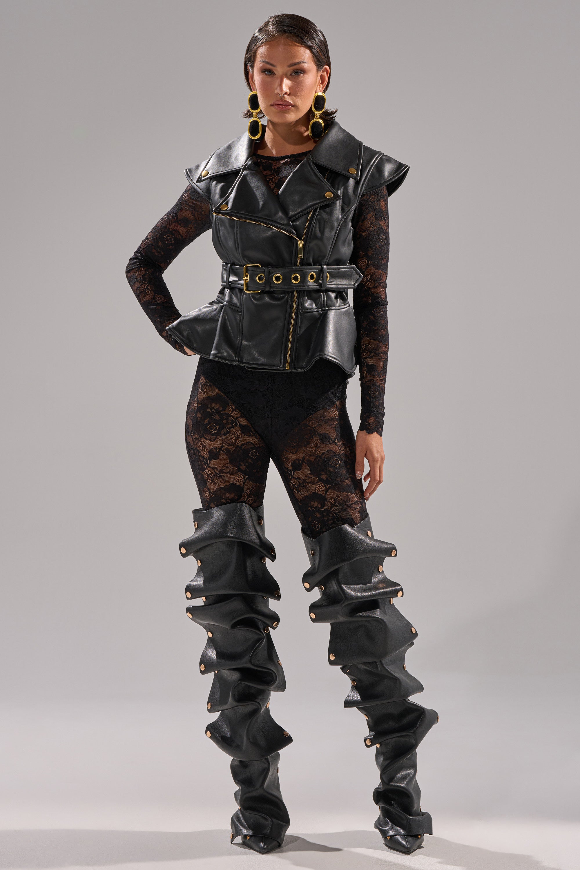 A woman poses in the PEP IN MY STEP LEATHER VEST, styled over a black lace bodysuit with matching oversized ruched leather boots and large gold statement earrings, standing against a plain gray background. 8