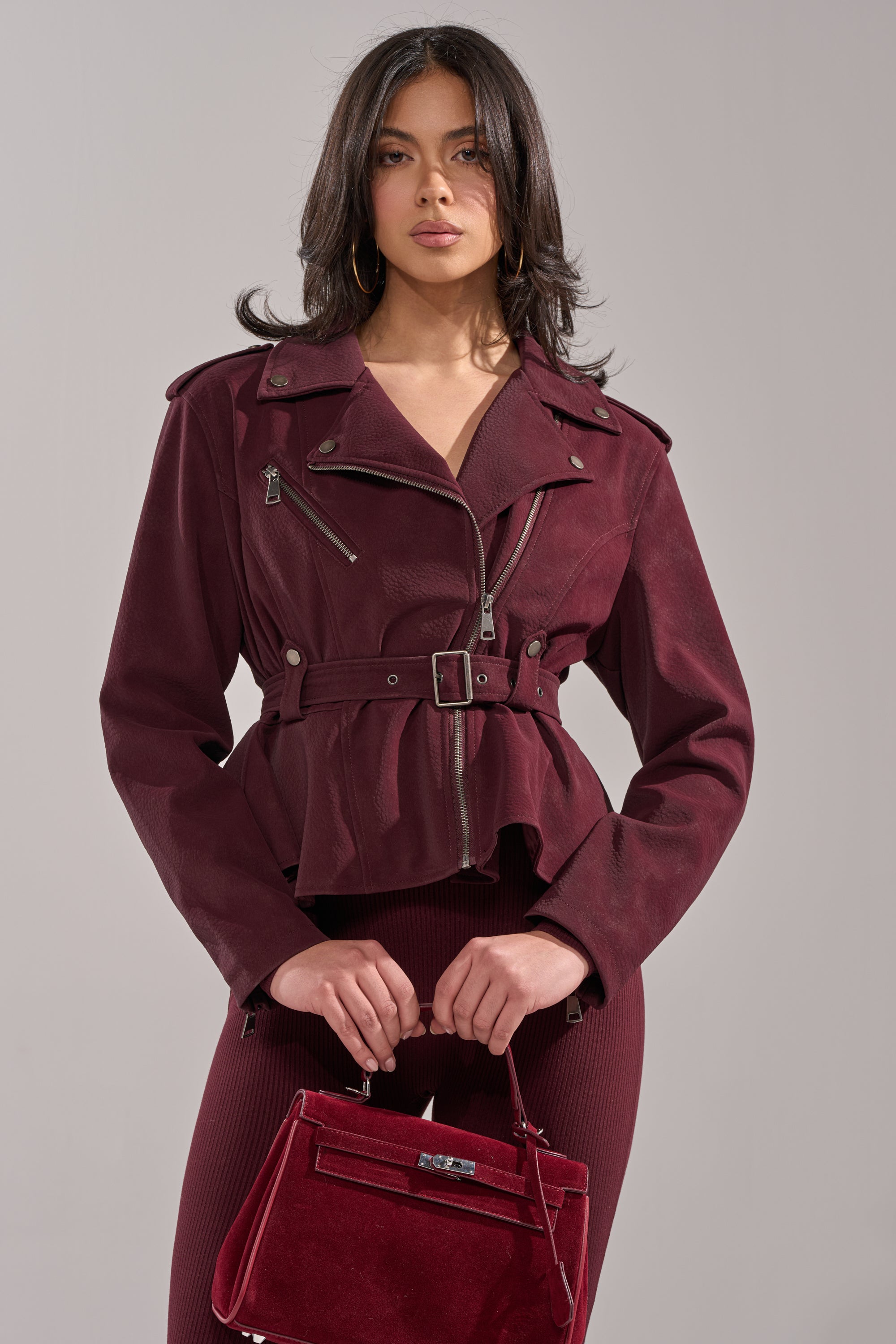 A woman confidently poses against a gray background in the DANI SUEDE EFFECT PEPLUM MOTO jacket over a matching ribbed dress, holding a burgundy handbag. 0