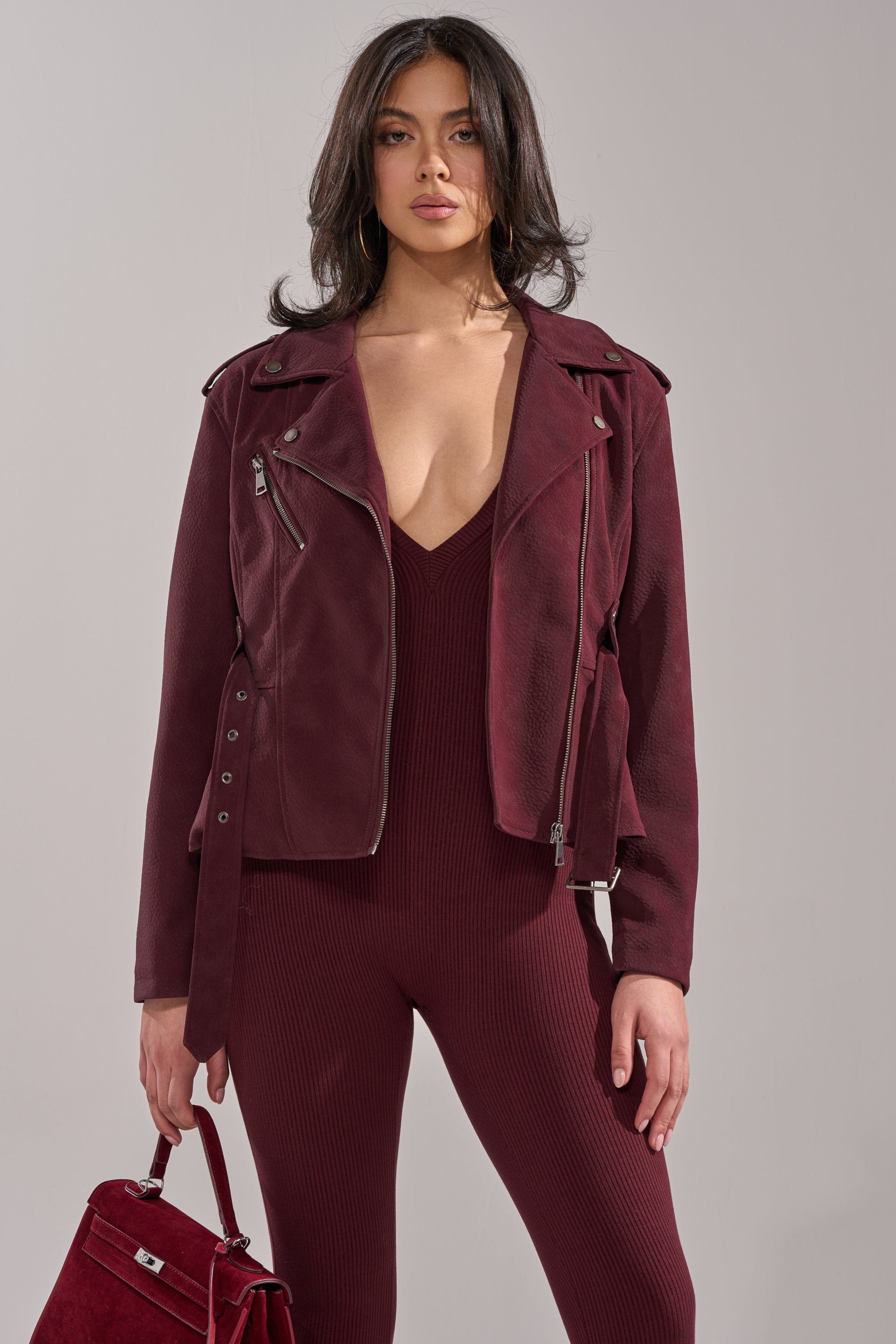 A woman with wavy dark hair wears the DANI SUEDE EFFECT PEPLUM MOTO jacket over a matching ribbed jumpsuit with a deep V-neckline, holding a burgundy handbag and standing against a neutral background. 2