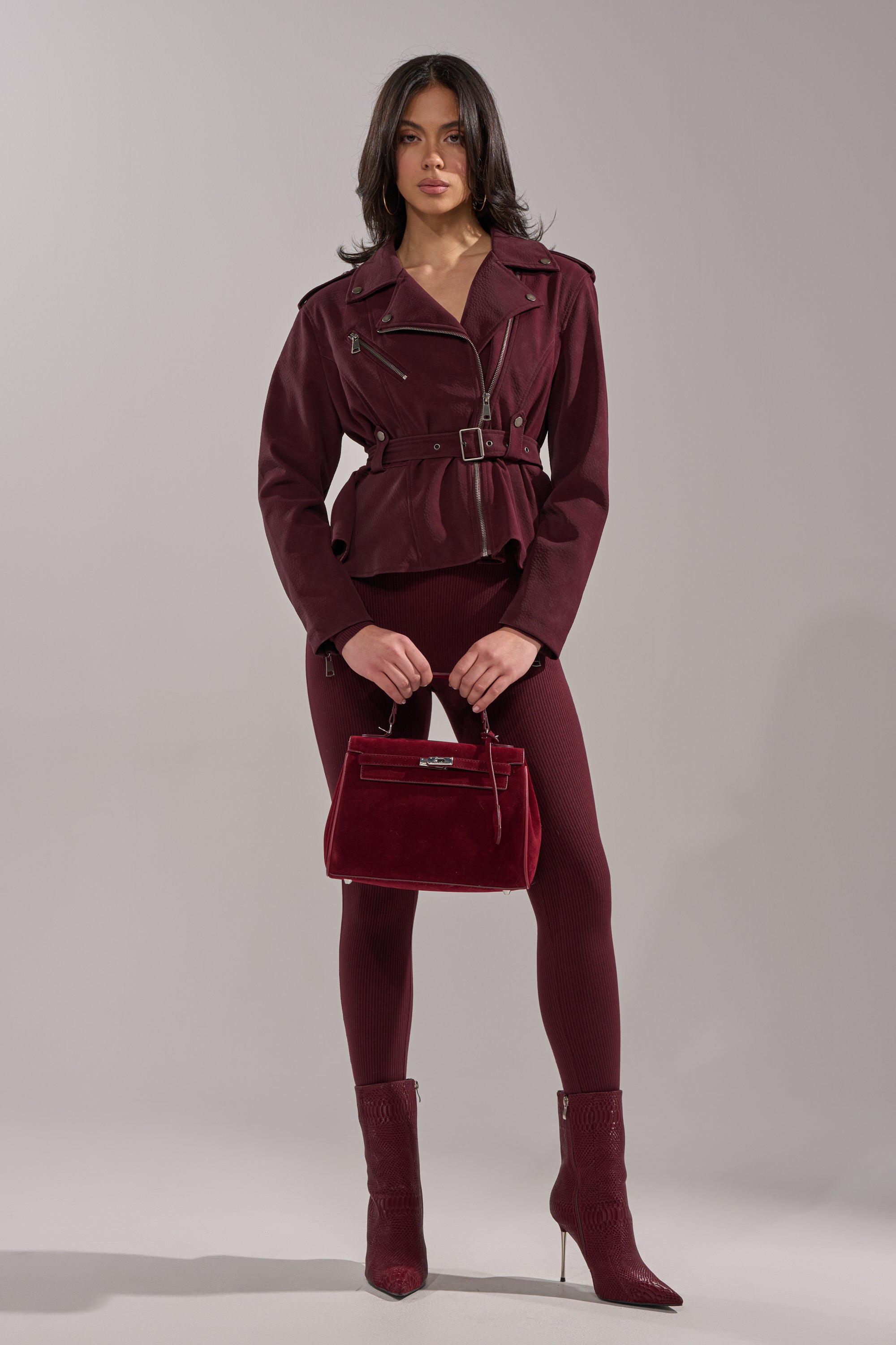 A woman with shoulder-length dark hair poses confidently against a gray background in the DANI SUEDE EFFECT PEPLUM MOTO jacket, styled with matching leggings, pointed-toe boots, and a red velvet handbag. 5