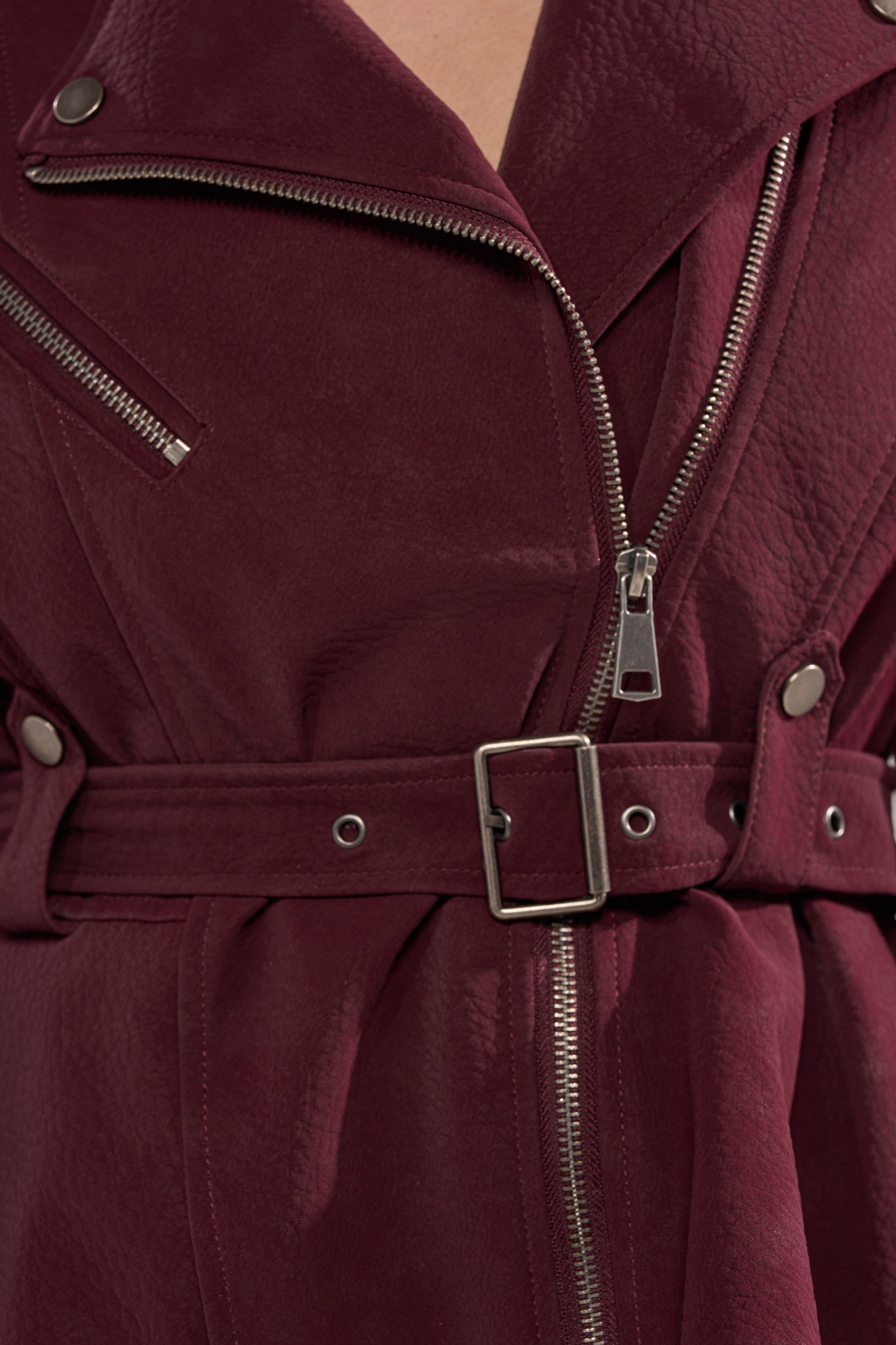 Close-up of the DANI SUEDE EFFECT PEPLUM MOTO—a burgundy faux leather jacket with silver zippers, belted waist, and snap buttons. This tailored biker blends edgy style with a modern peplum silhouette. 6