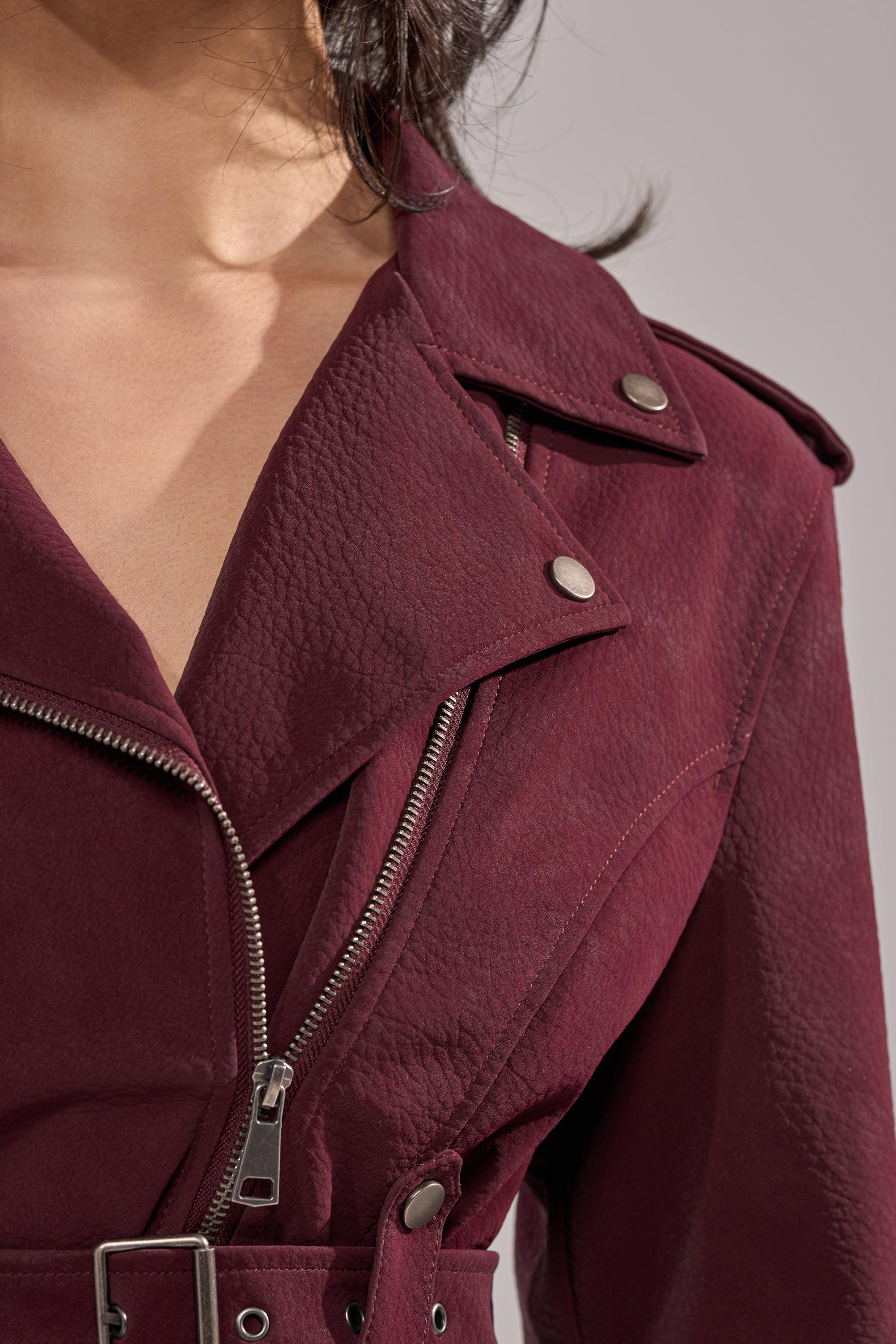 A close-up of a person wearing the DANI SUEDE EFFECT PEPLUM MOTO, a textured burgundy biker jacket with silver snap buttons, zipper, belted waist, and wide collar. The person's neck and part of their chin are visible. 7