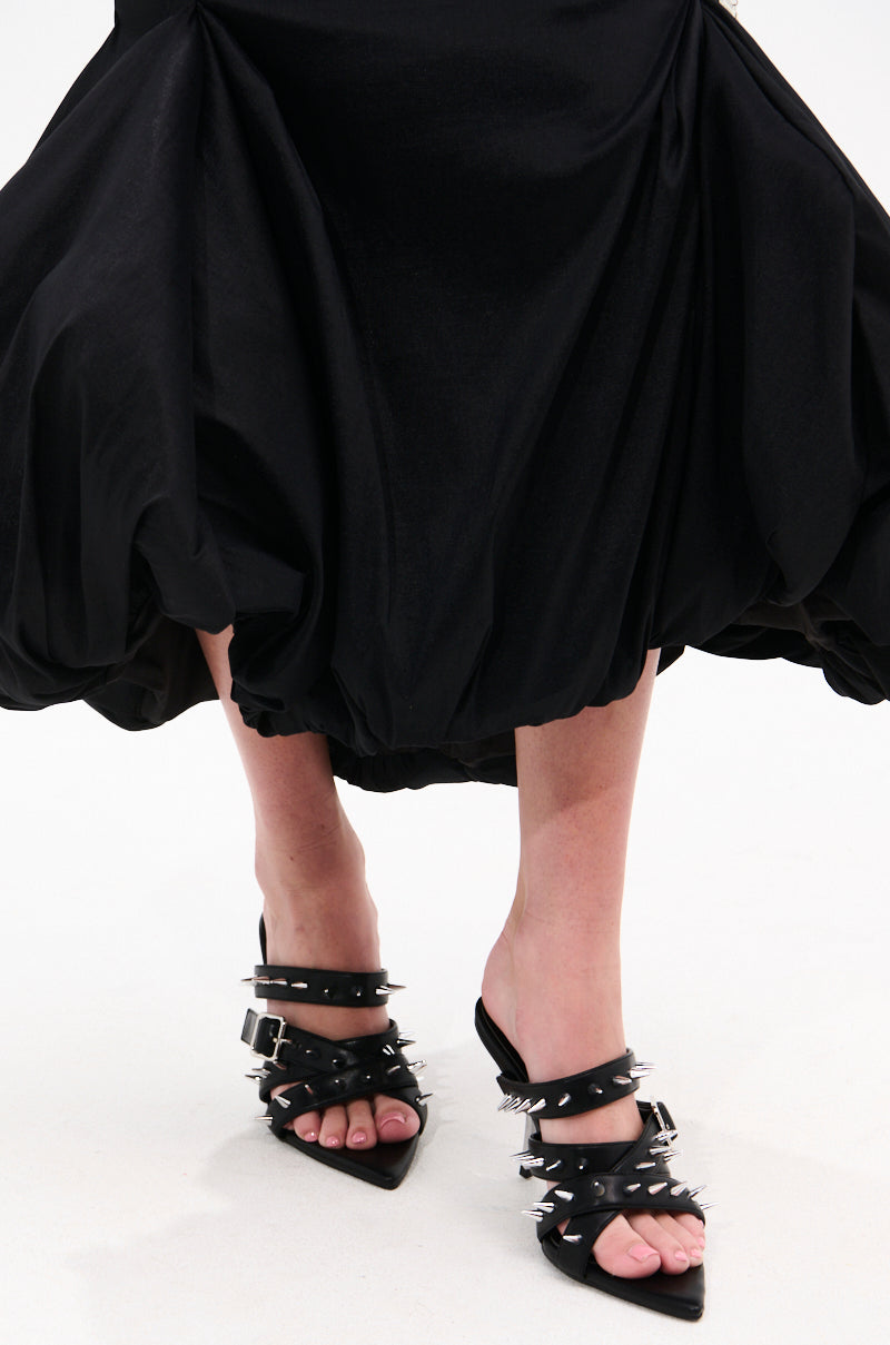 A person in a voluminous black skirt and the AZALEA WANG PEREGRINE BLACK STUD AND SPIKE MULE SANDAL with stiletto heels stands on a white background. 1