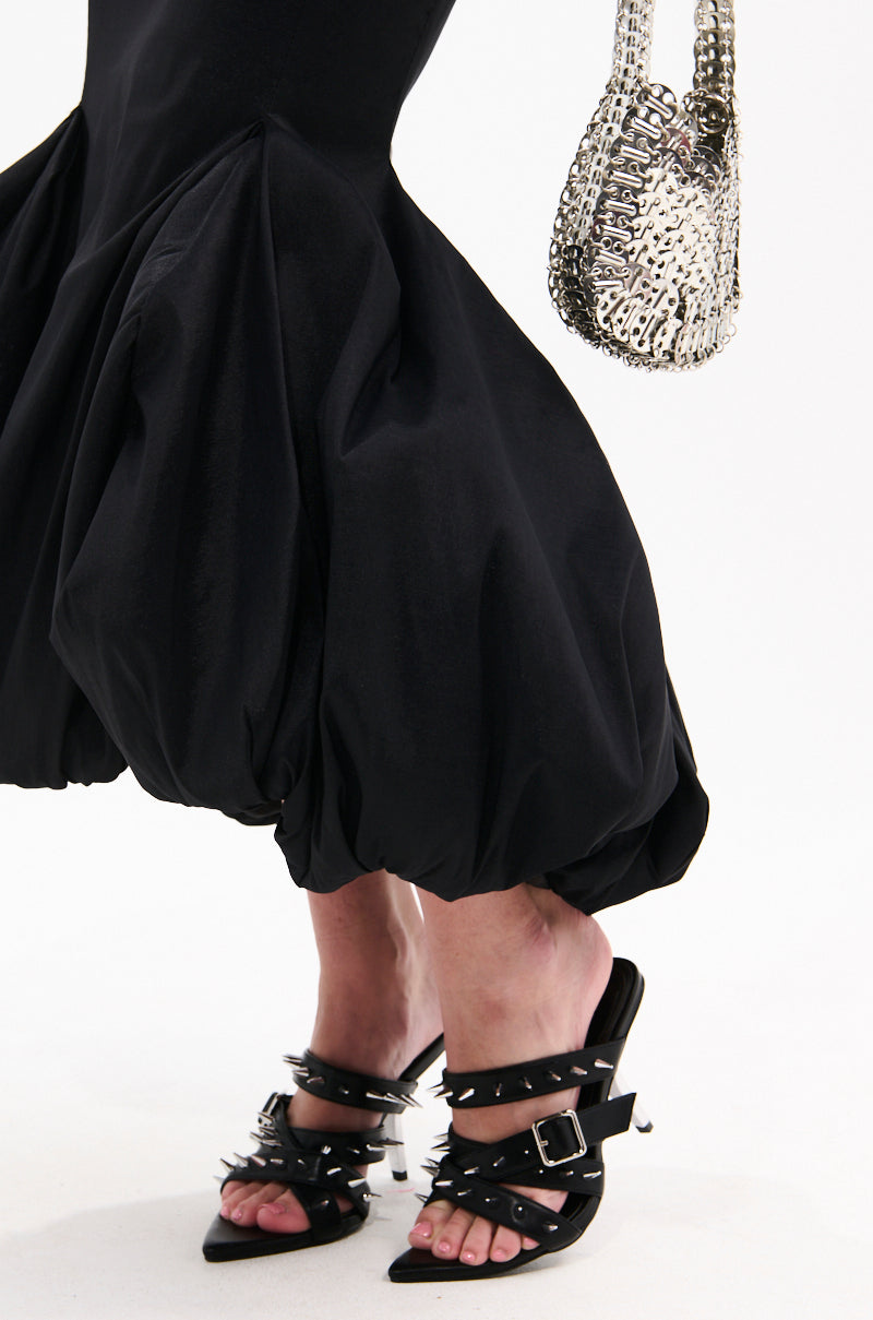 A person wears a black, voluminous faux leather balloon-hem skirt, AZALEA WANG PEREGRINE BLACK STUD AND SPIKE MULE SANDALS, and holds a silver chainmail-style handbag against a white background. 2