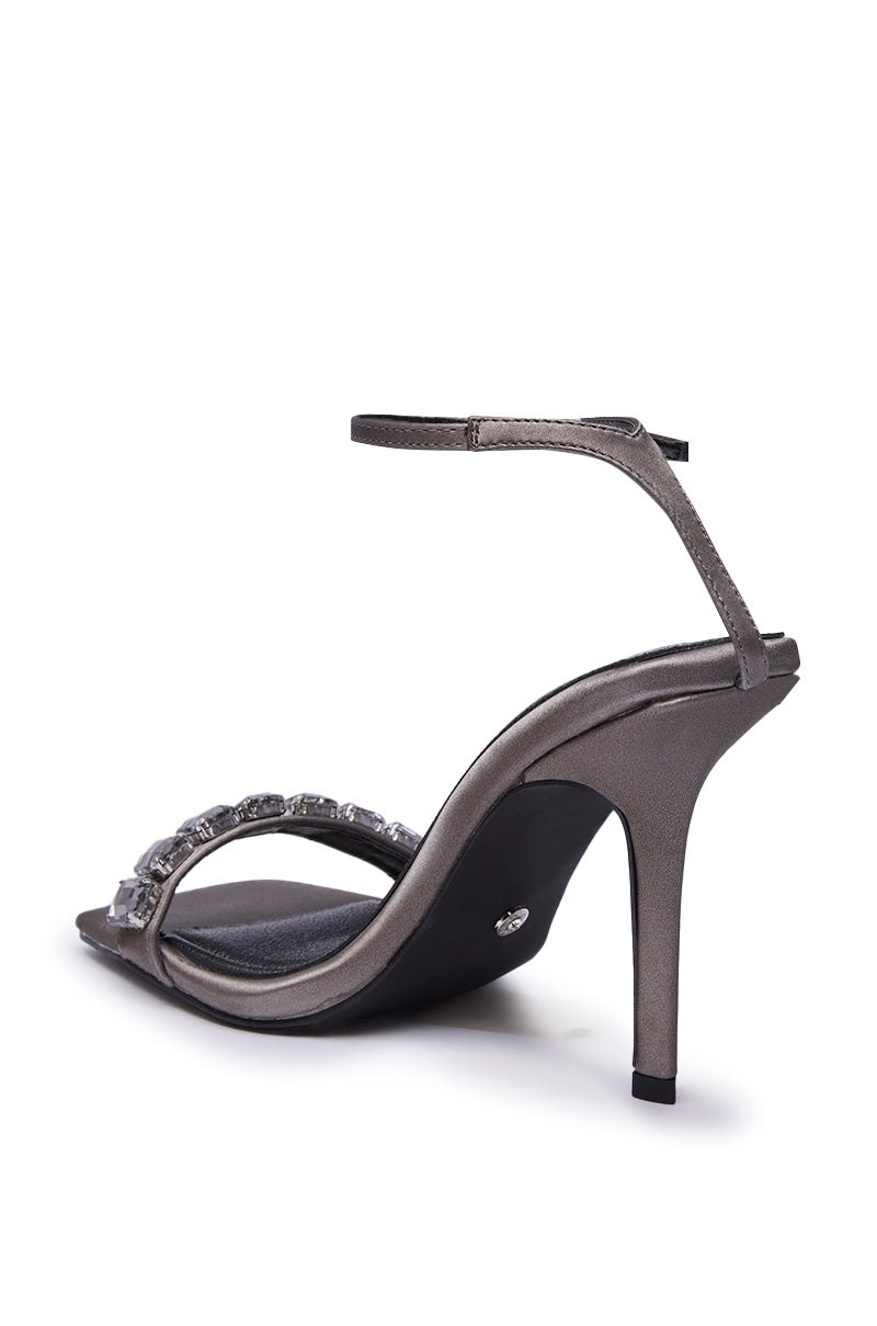 The AZALEA WANG PERIIS GREY RHINESTONE SANDAL features a metallic gray faux leather build, stiletto heel, ankle strap, and gemstone-embellished front strap, shown from a rear diagonal angle on a white background.