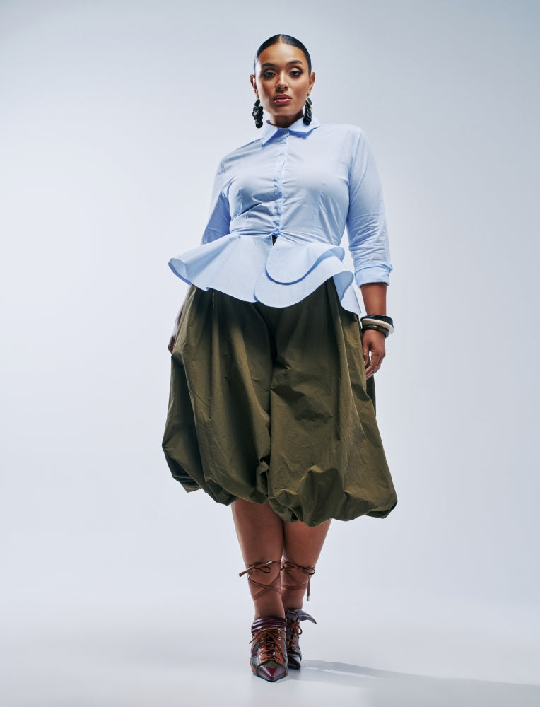 A model stands against a light gray studio background wearing a light blue long-sleeve peplum-style blouse tucked into olive green wide-leg pants, paired with brown lace-up ankle boots.